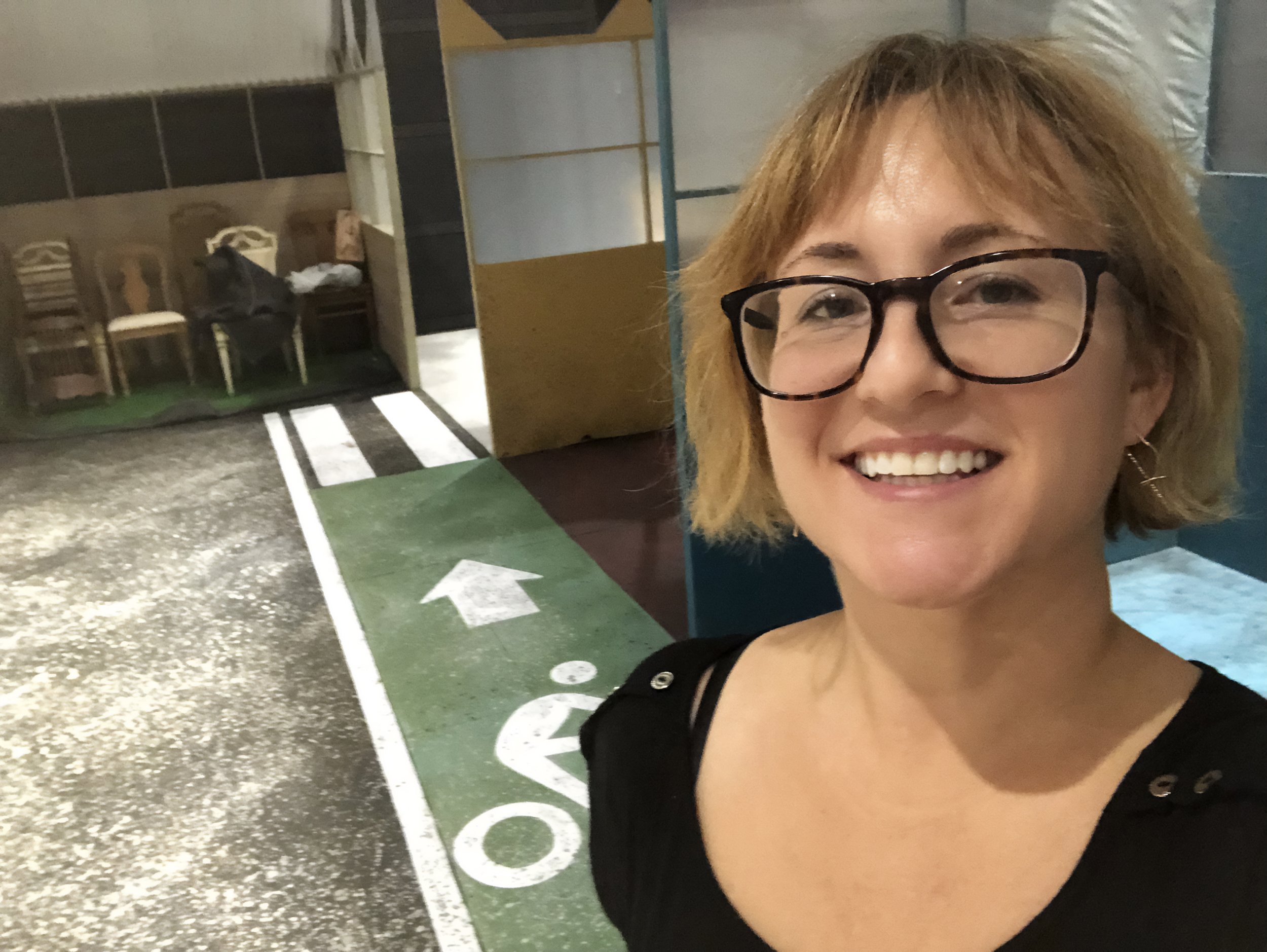 Smiling woman with short blonde hair and glasses in an indoor setting, with a bicycle lane marking on the ground and chairs in the background.