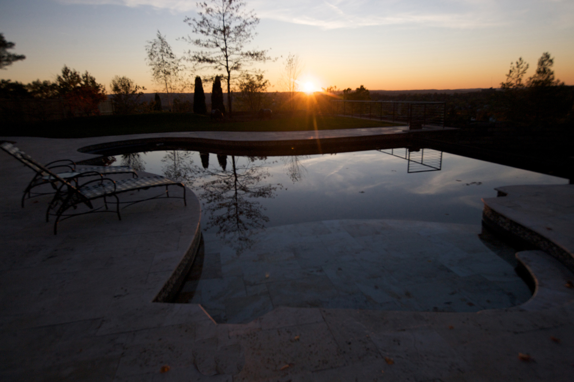 This property is situated in a residential community overlooking the City of Syracuse, New York. Perched high on a hill, the site has wonderful sunsets and magnificent views across the city. A steeply graded slope was converted into two level terraces, encouraging outdoor entertainment and a gracious garden environment. The top terrace includes a ‘free form’ Gunite swimming pool with a vanishing edge hovering over the landscape. This edge drops 9ft to create a ‘water wall’ over natural stone into a small pool on the lower lawn terrace. The lower lawn terrace was designed for sports and play, and it’s an ideal location for a tent for during outdoor functions. We elected to plant some large specimen trees and shrubs for instantaneous visual impact; otherwise, native plant materials were used throughout the site to accommodate existing site conditions and reduce landscape maintenance. A J Miller Landscape Architecture handled the lighting design of this project. Brass cast light fixtures were chosen for durability and water resistance. Indirect lighting principals were employed: light fixtures are concealed and blended into the hardscape and gardens. We were instrumental in advising our client to repaint the existing home, from white and grey to a warm beige with green trim highlights. In addition, the client installed a new brown roof with copper gutters. This ‘anchored’ the house to the site and complemented the hard landscape materials, which helped with the scale of the project by creating a more inviting atmosphere. 