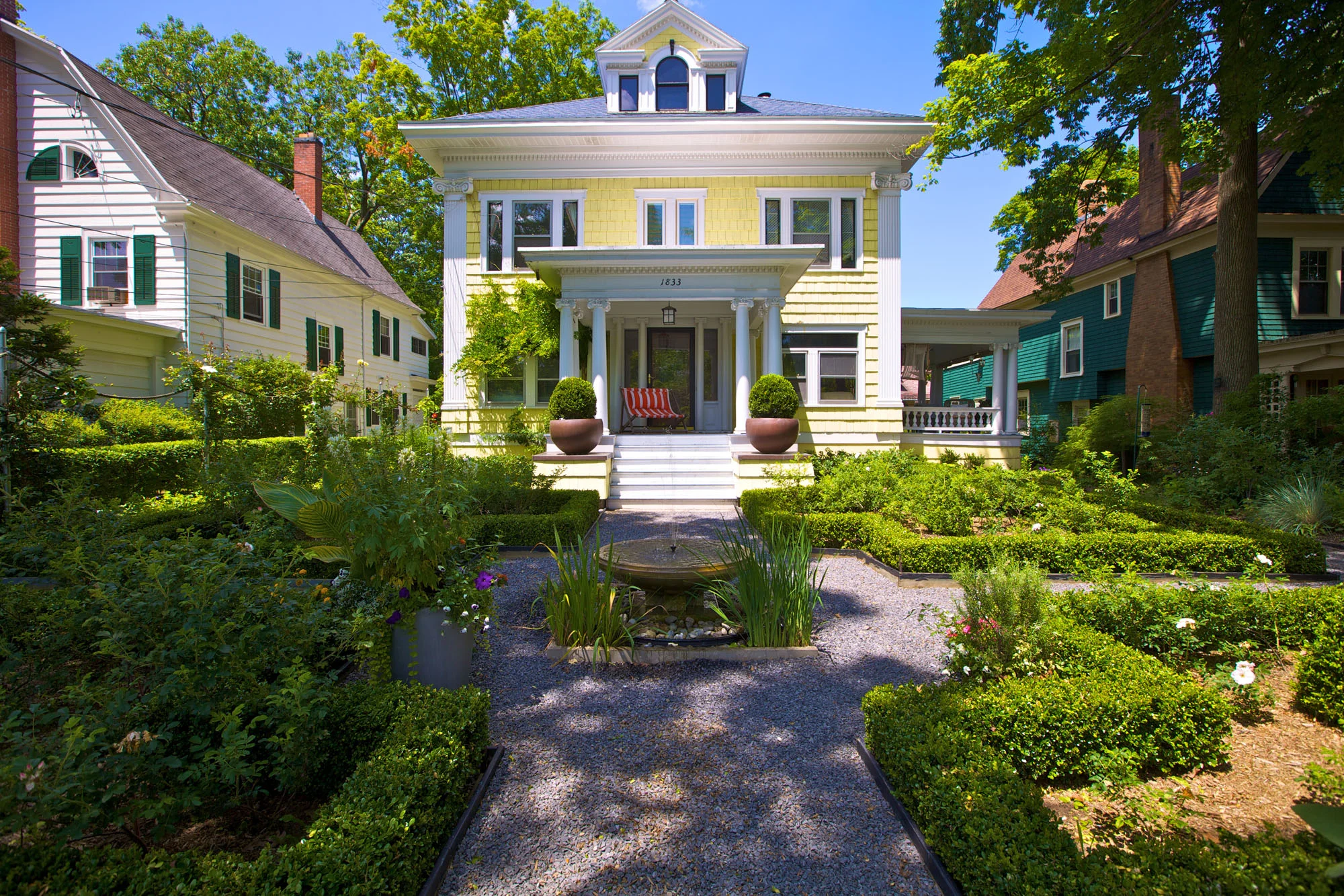 The home and office of A J Miller Landscape Architecture is situated atop a hill in the Eastwood neighborhood of Syracuse, New York. Built in the 1911 during the Arts & Crafts period, the home is considered an American Foursquare Colonial. The elevated placement of the house provides an ideal setting for a French parterre garden, a landscape meant to link the architecture of the home with its gardens by promoting a visual dialog between the natural and built environment. Drawing from the Arts & Crafts design philosophy of the house, the Foursquare pattern of the garden echoes the motif of stained glass windows overlooking the parterre. Boxwoods frame the focal point of the garden: a limestone fountain whose bubbling, clear water is a favorite drinking spot for songbirds. In the summer this water feature is the life of the garden. The garden’s color palette is composed of purples, whites, yellows, silvers, and blacks; a delightful contrast with the home’s yellow exterior and grey roof. The rear garden is a shade garden facing North, many shade plants surround a gravel terrace. A clump Bamboo grove is on two sides this provided shelter from prevailing winds and provides seclusion for a sectional seating area around a copper fire pit.