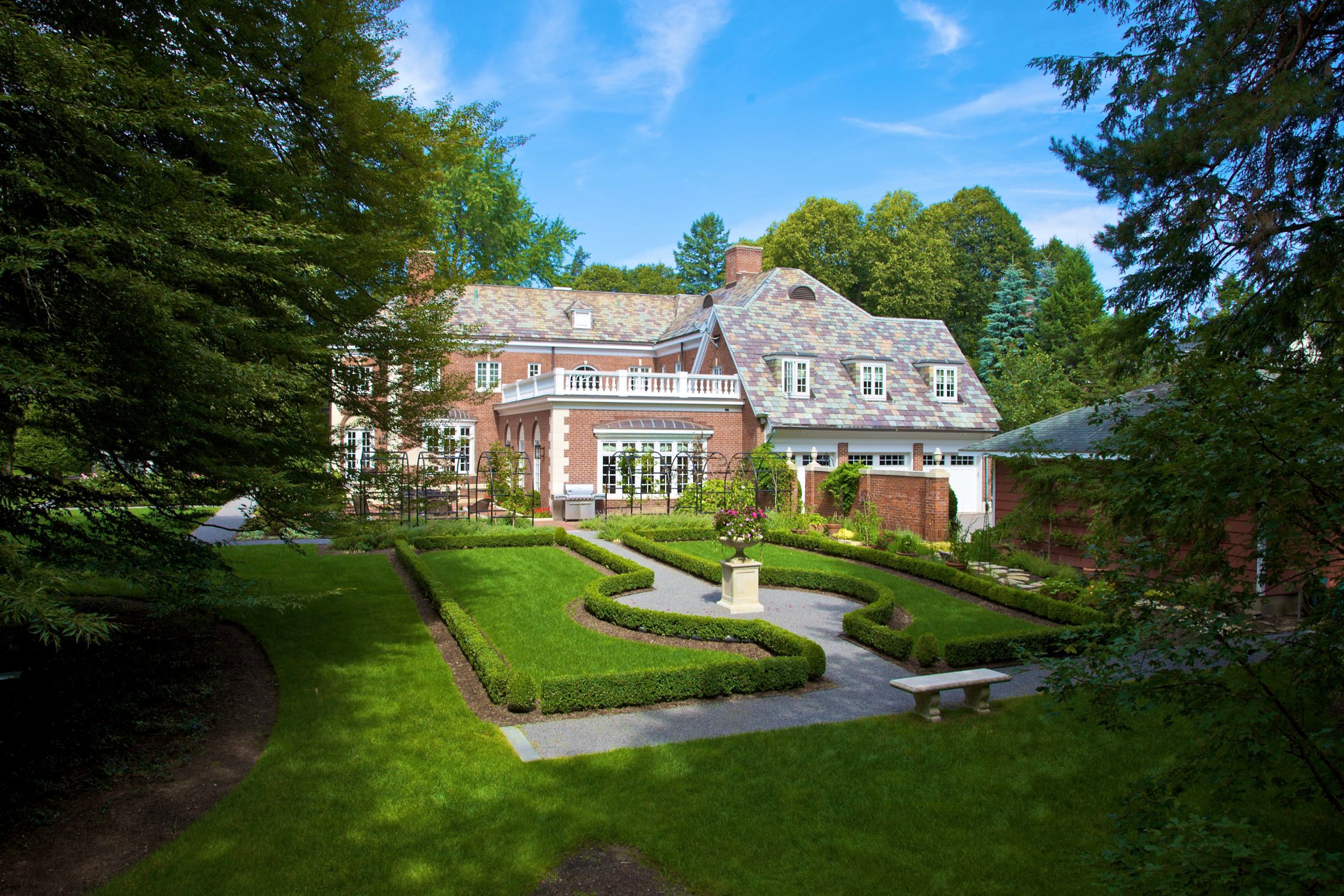 This property is situated in a residential community just outside the city of Rochester, New York. The home is on a large double city lot in a distinctive neighborhood, whose architectural character spans from 1900 to the present day. A.J. Miller Landscape Architecture designed the driveway for guest drop-off at the grand entrance of the home. Some visitors are permitted to park in the half circle driveway, which is lined with a drivable, or “rolled” curb. This hardscape design element deliniates allowable driving areas, with room to accommodate guests. Brick pavers line the walkways, encouraging a visual dialog between the house and landscape . The paths are outlined with 8” wide flame finished custom granite accent strips. A combination of of running bound with center panels in basket weave bond creates an elegant and interesting paving pattern. A.J. Miller Landscape Architecture wanted to create a garden to reflect the sophistication of the house and surrounding neighborhood. The existing gardens had been overgrown for many years, and were dominated by an unimaginative lawn and dreary planting beds. The clients wanted a garden reminiscent of what they had seen on visits to England and Western Europe. In particular, it was important that the landscape could be used for the entertainment and enjoyment of their young children and family friends. The planting design required the removal of several overgrown trees; this new light and open space allowed for the creation of formal outdoor garden rooms. A wrought iron metal archway leads the visitor from the parking garage and forecourt through the herb garden, which is used extensively by the client for culinary applications. Secondary garden pathways lined with gravel branch off in regular intervals from the main passage. A.J. Miller Landscape Architecture devised a large brick terrace for outdoor dining and relaxation; this area is bordered by substantial limestone trough planters. Every season the planters are adorned with lavish horticultural arrangements. A vista overlooking a formal parterre can be enjoyed from a bench at the edge of the terrace, which rests upon an elegant plinth and large limestone urn. The parterre is a garden room whose borders are defined by low boxwood hedges. A second formal garden room is located on the side of the house near a small private seating terrace– an extension of the interior living room. Here we designed a colorful ‘rose & peony’ garden edged with Boxwoods, with a pyramid-shaped Buxus marking each corner. The garden is framed from the street with a row of mature Hornbeams that were to eventually trained as a ‘pleached hedge”. Visitors can follow pathways from the terrace to Great Lawn under an archway planted with purple and copper beech climbers. Scented perennials anchor the space under the arch. The Great Lawn was established by removing some overgrown trees while protecting an ancient English Beech in the center of the landscape. With this tree canopy gone we gently leveled the lawn and re-laid new sod. A woodland border of specimen trees, shrubs, and shade-loving perennials provides a multi-layered boundary on one side of the space.