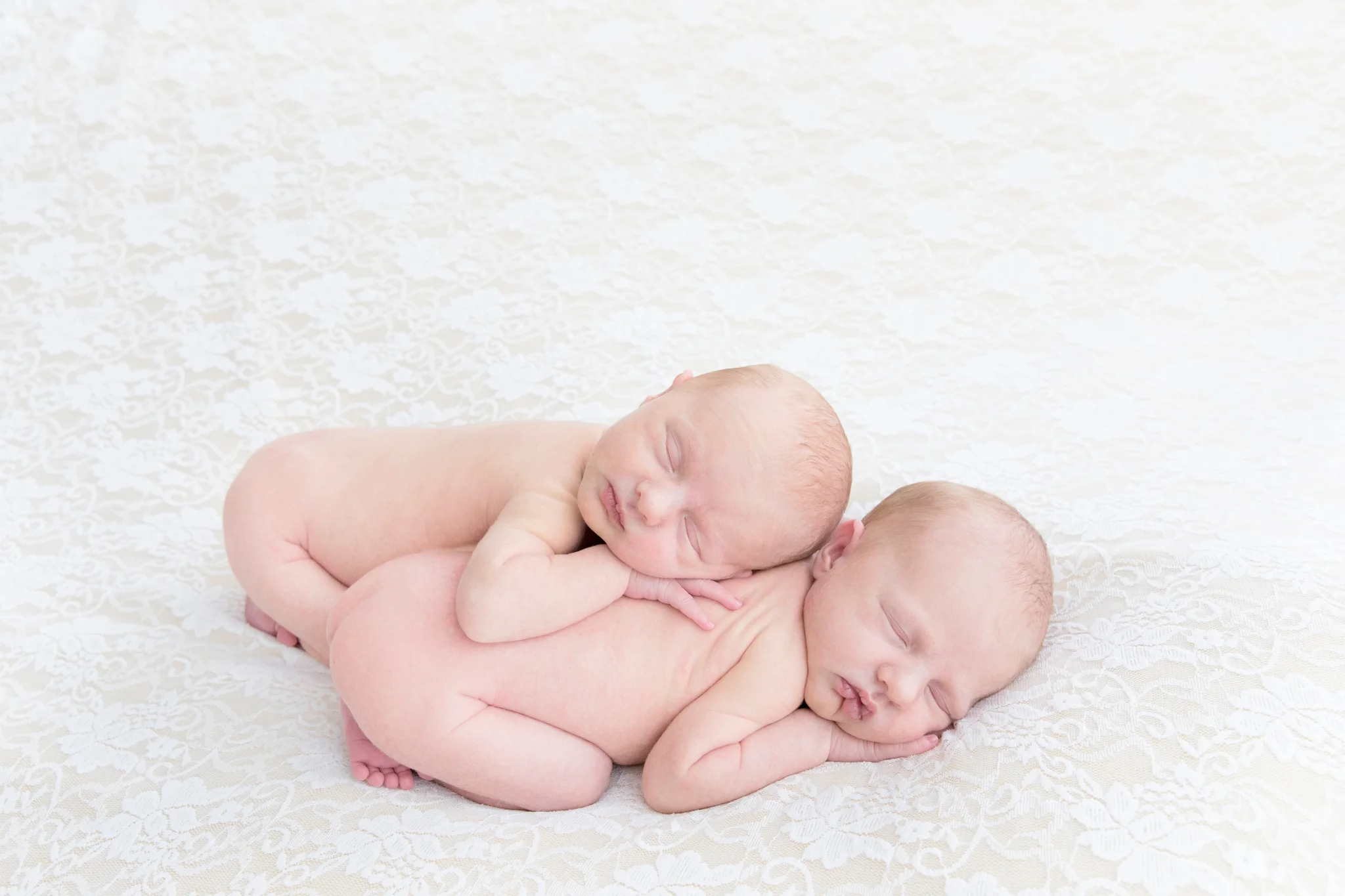 Newborn twin girls cuddling on a white and cream lace blanket.