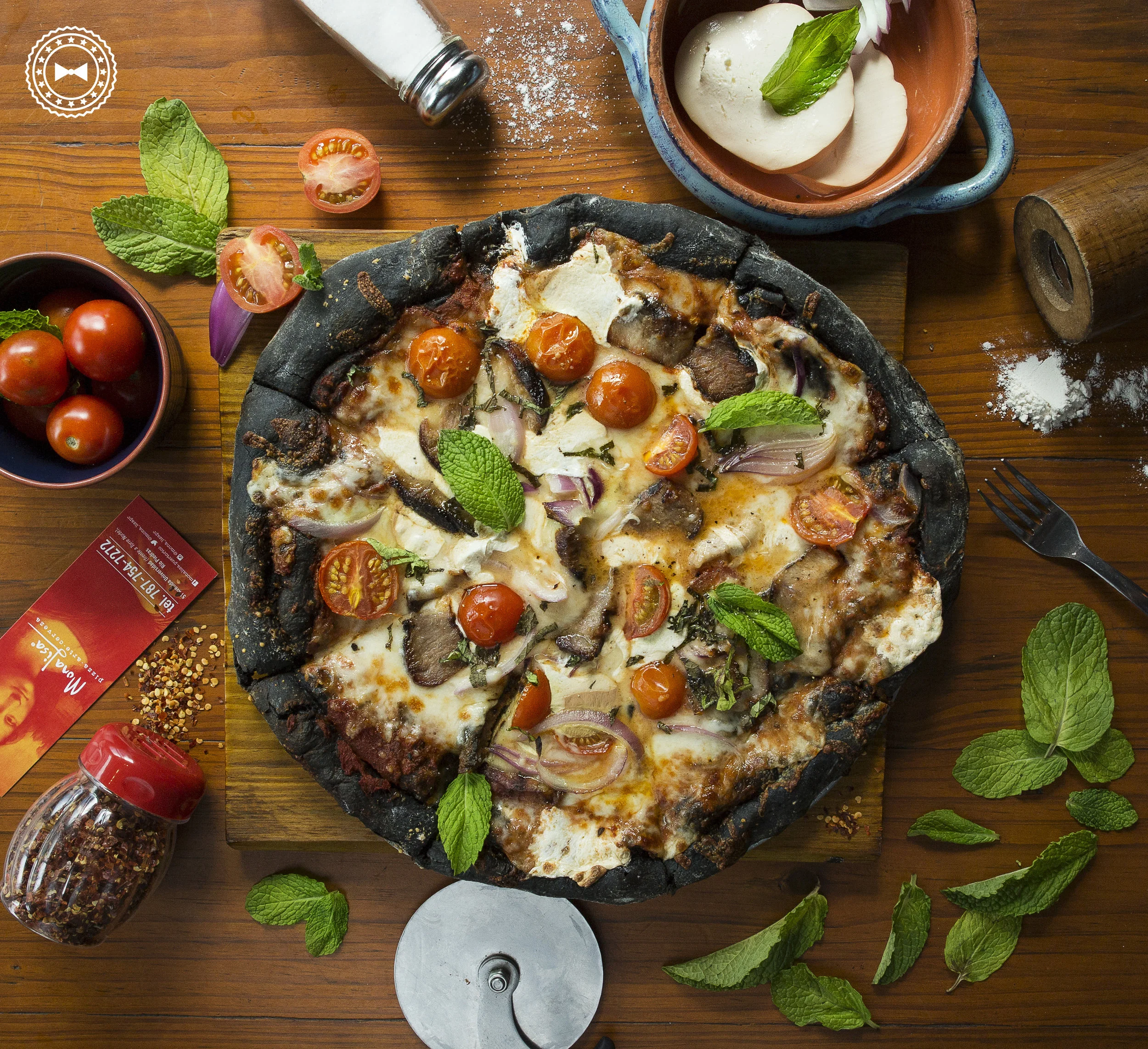 Pizza Negra (masa negra a base de tinta de calamar, salsa pomodoro, queso mozzarella ahumado, cerdo a la parrilla, cherry tomatoes, cebolla lila y hojas de menta), de Mona Lisa 