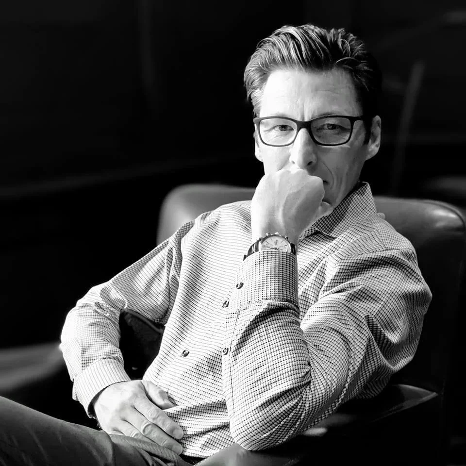 A black and white photo of a man sitting in a chair, wearing glasses and a checkered shirt. He has his hand covering his mouth and is looking directly at the camera.