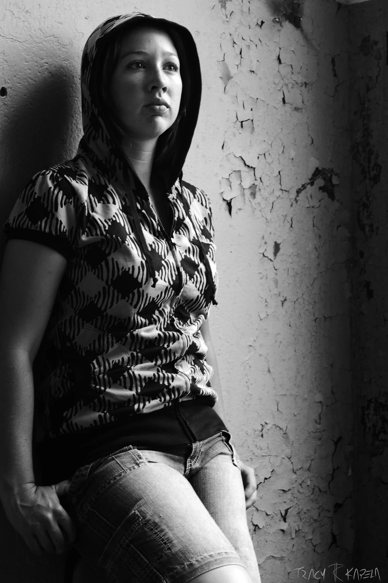 A black and white photo of a young woman wearing a patterned hoodie and denim shorts, sitting against a textured, cracked wall, looking into the distance with a contemplative expression.