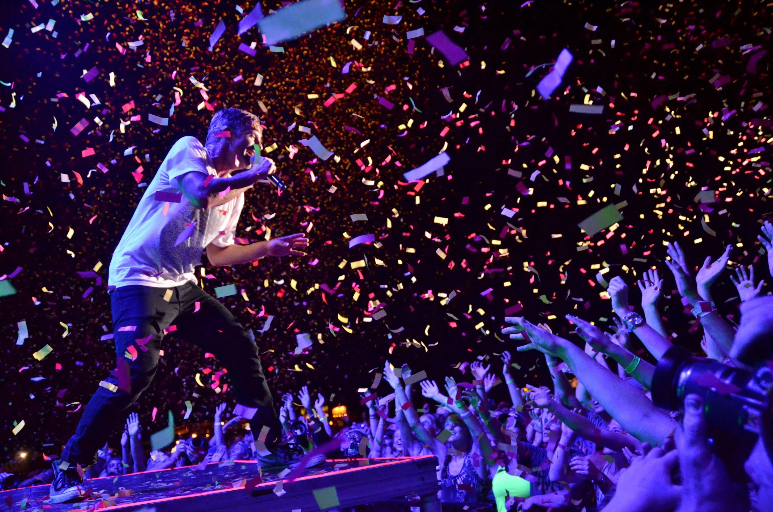 A performer singing on stage with confetti falling, reaching out to an enthusiastic crowd with hands raised.