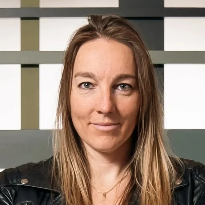 Portrait of a woman with long, wavy light brown hair, wearing a black leather jacket and a gold necklace, standing in front of a modern geometric background.