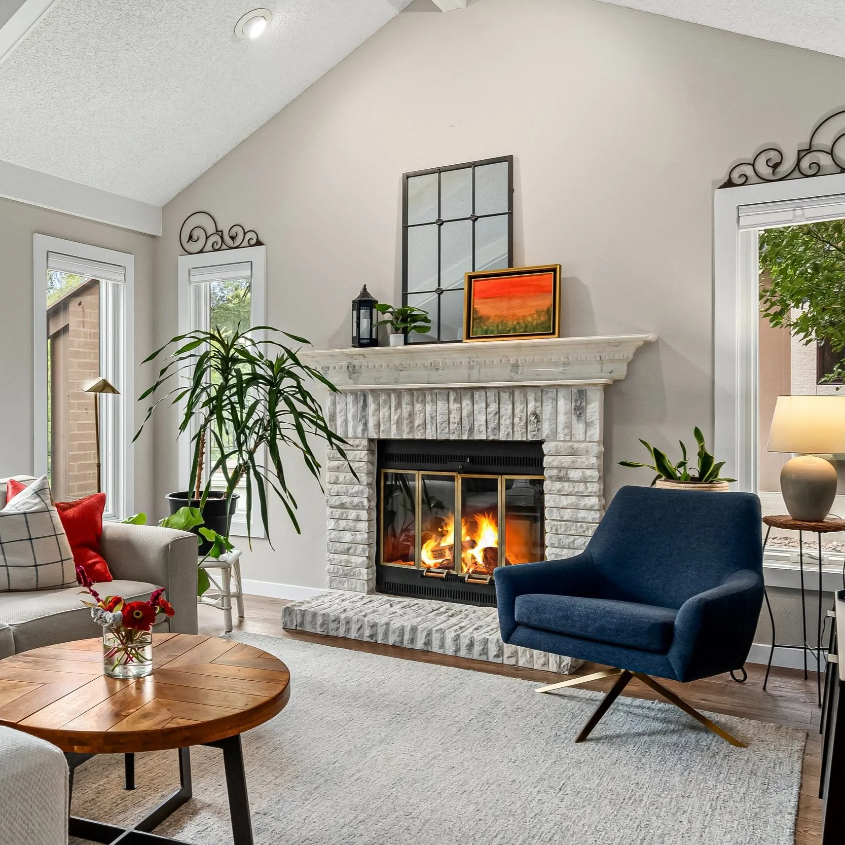 Living room with a lit fireplace, blue lounge chair, beige sofa with pillows, wooden coffee table with a glass vase of red flowers, large houseplants, and windows with white trim and decorative ironwork over the window frames.
