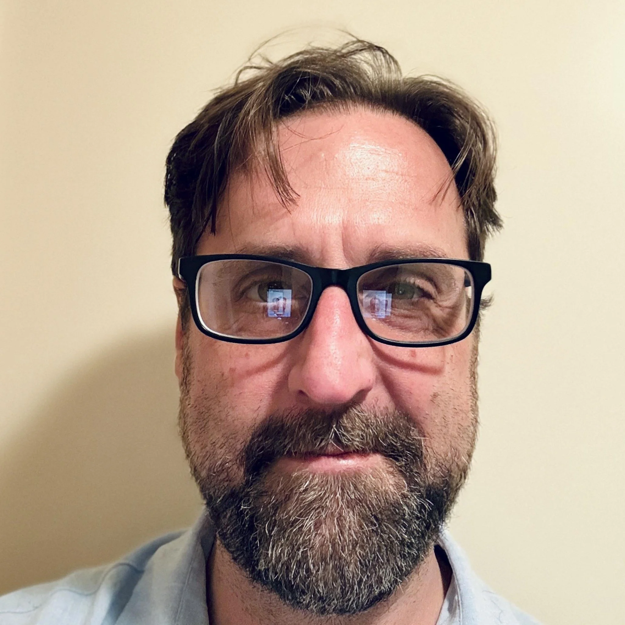 Close-up photo of a middle-aged man with glasses, a beard, and brown hair, standing against a plain light-colored wall.