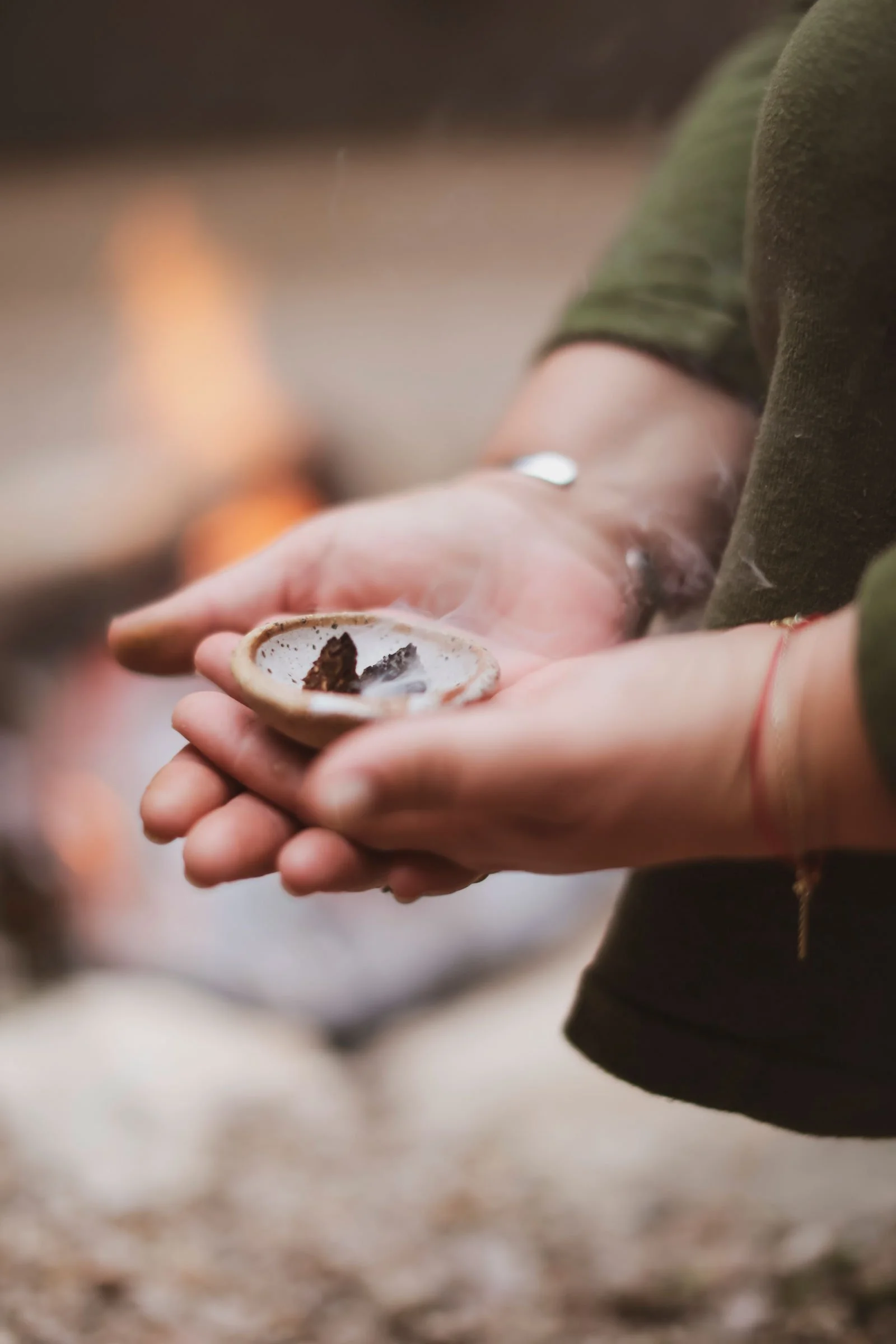 Person holding a small bowl with burning herbs or incense, with visible smoke, in an outdoor setting.