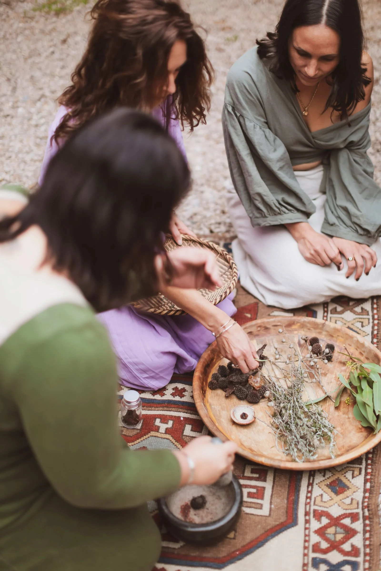 Three women sitting on a patterned rug outdoors, engaged in a natural crafting activity with dried herbs, pinecones, and other natural materials on a wooden tray.