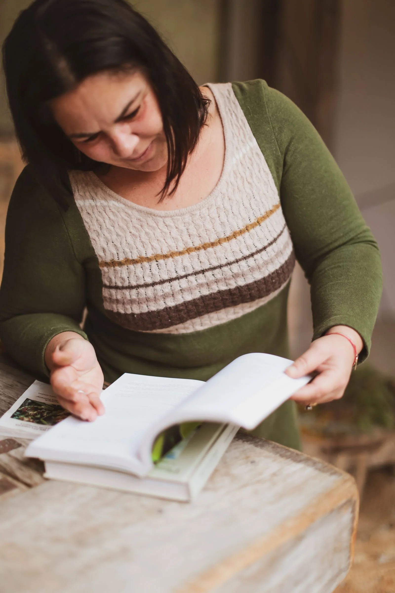 A woman with dark hair wearing a green sweater with a beige, brown, and khaki pattern reads a book at a rustic wooden table.