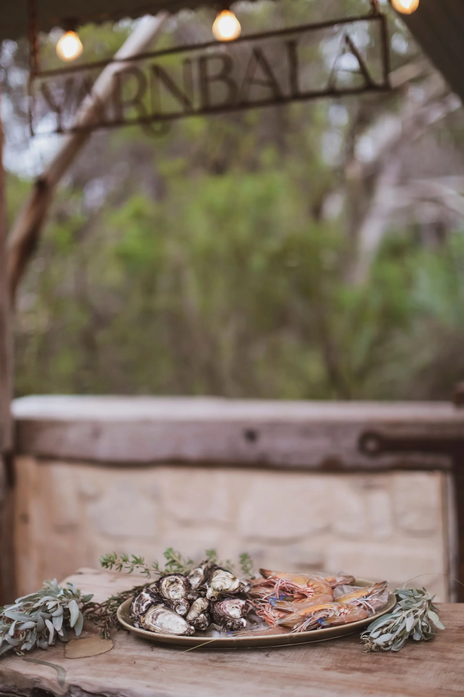 A rustic outdoor seafood platter with oysters and shrimp on a wooden table, decorated with green leafy herbs, in front of a stone wall and blurred trees."RNBALAK" sign hanging above with string lights.