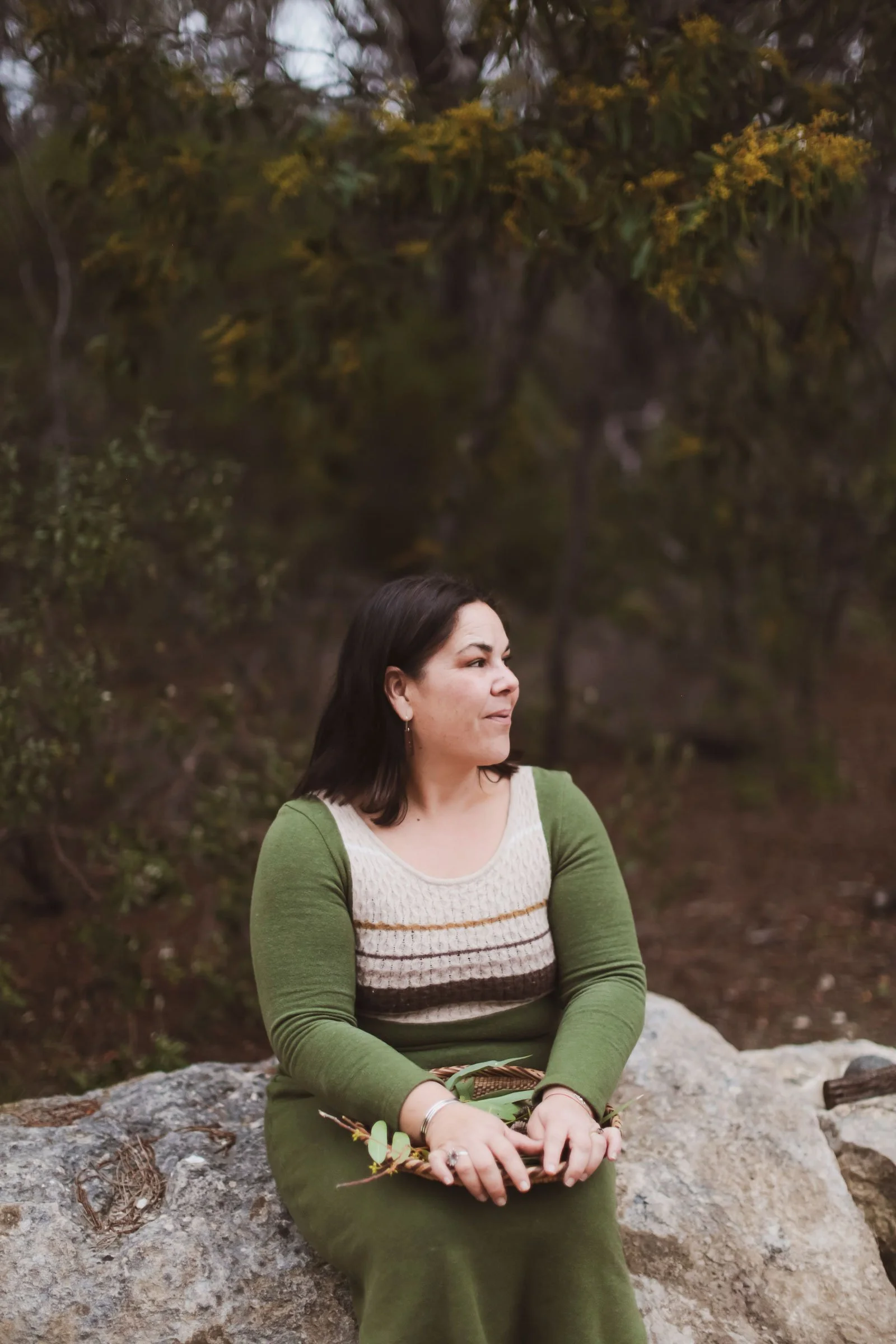 A woman in a green long-sleeved dress sitting outdoors on a large rock with trees and foliage in the background.