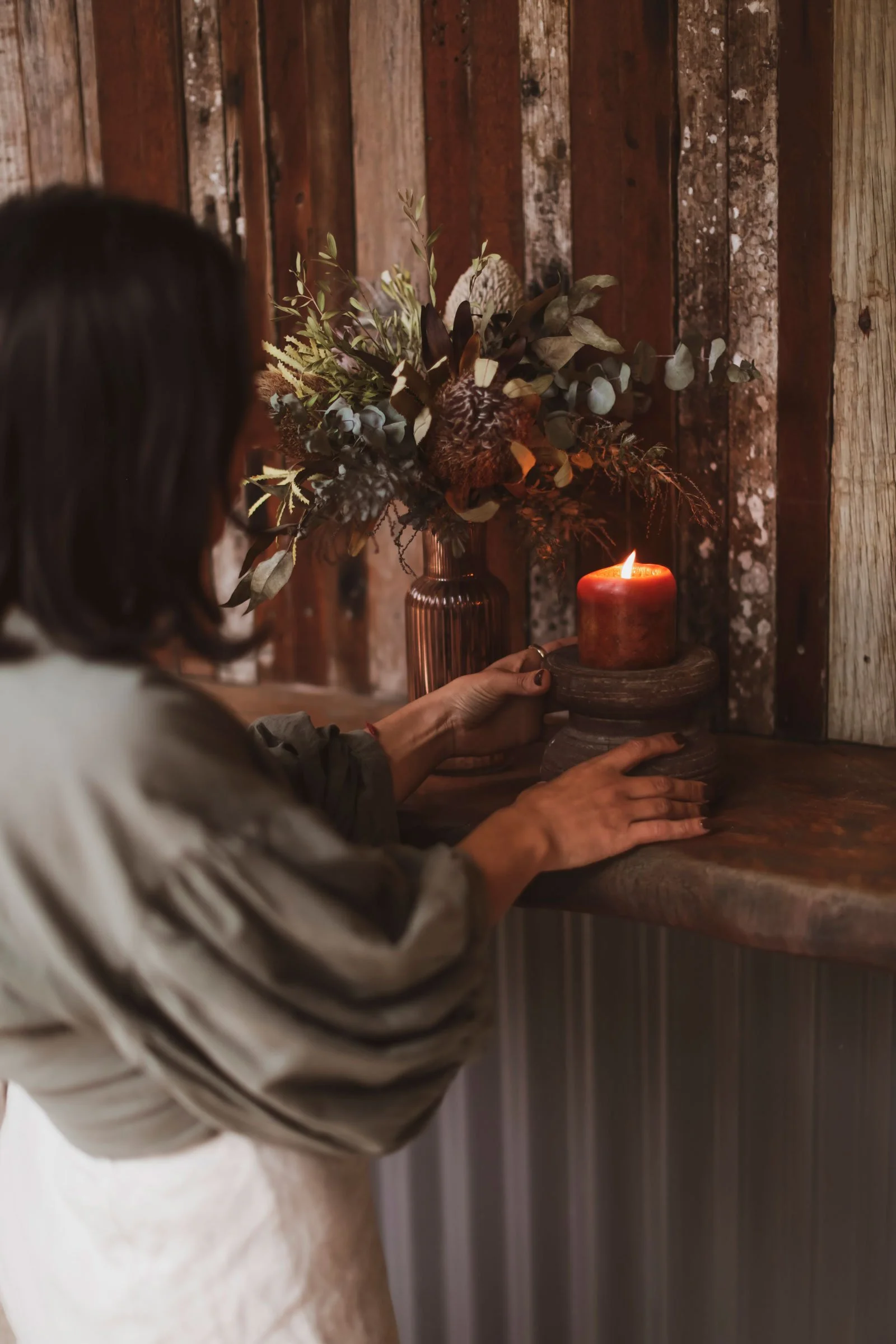 A person with dark hair lighting a red candle on a rustic wooden surface, with a bouquet of flowers in a vase in the background, against a wooden wall.