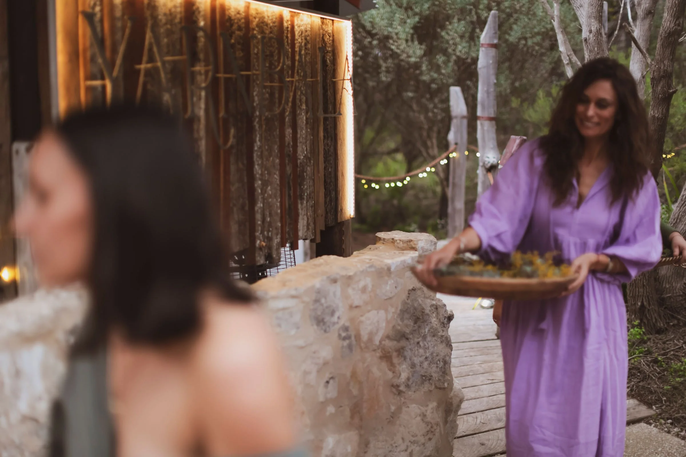 A woman in a purple dress serving food at an outdoor gathering, with blurred guests in the foreground and trees with string lights in the background.