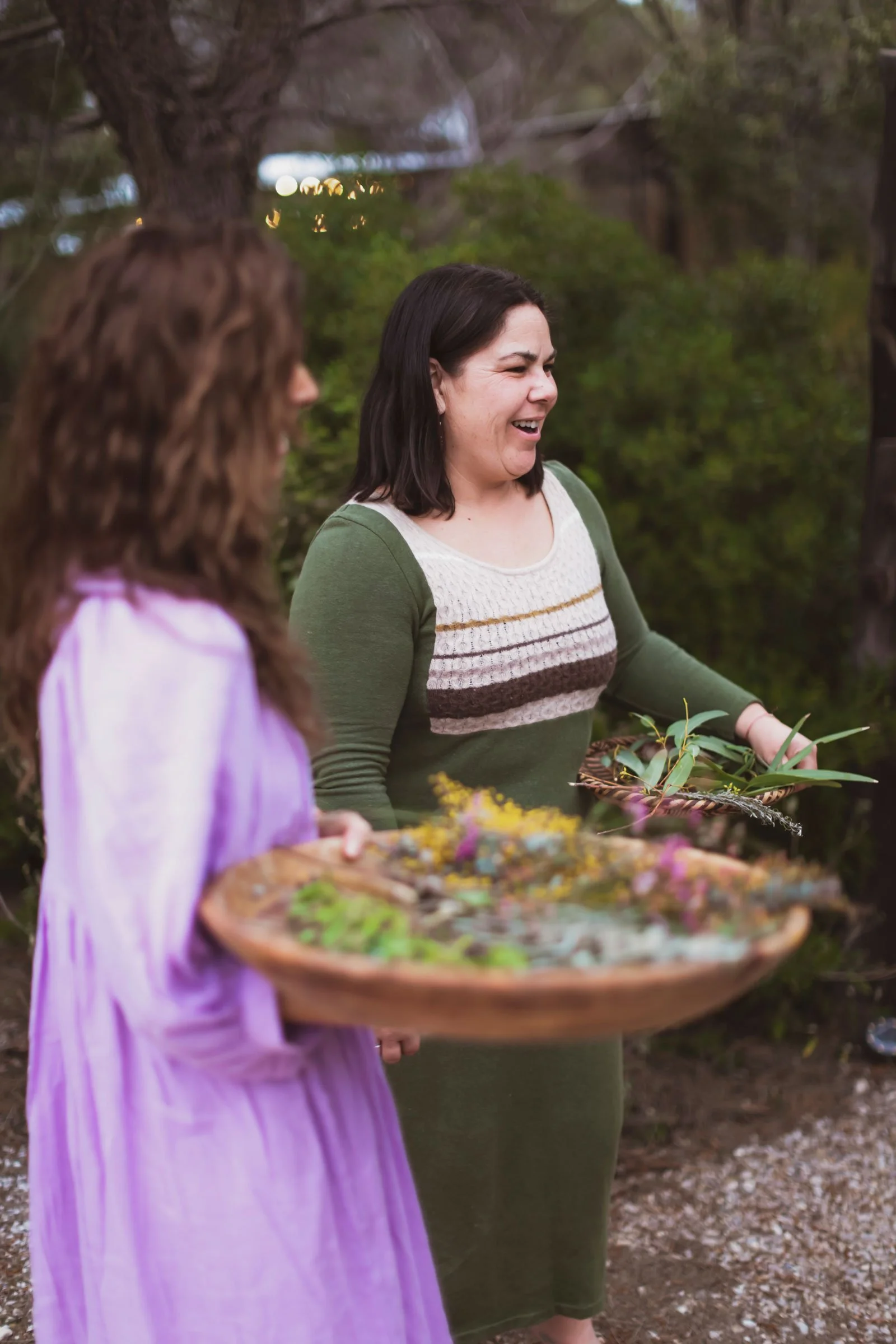Two women outdoors, one wearing a green and white sweater, holding a basket of greenery, and the other in a purple dress, holding a large tray of flowers and leaves, smiling and talking.