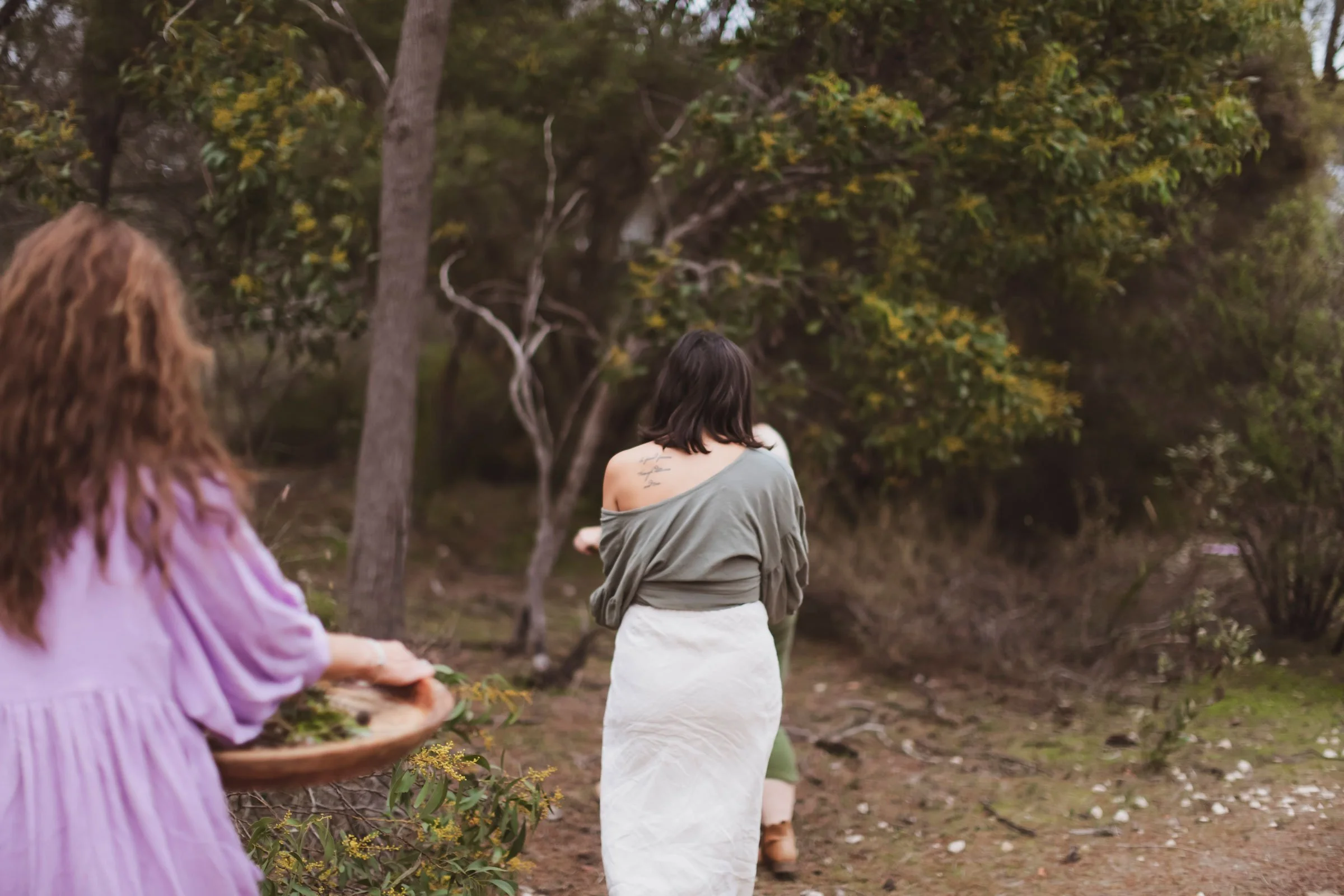 A woman with dark hair wearing a loose green top and a white skirt is walking away through a wooded area with trees and shrubs. A person in a purple dress is partially visible in the foreground, holding a basket of greenery.