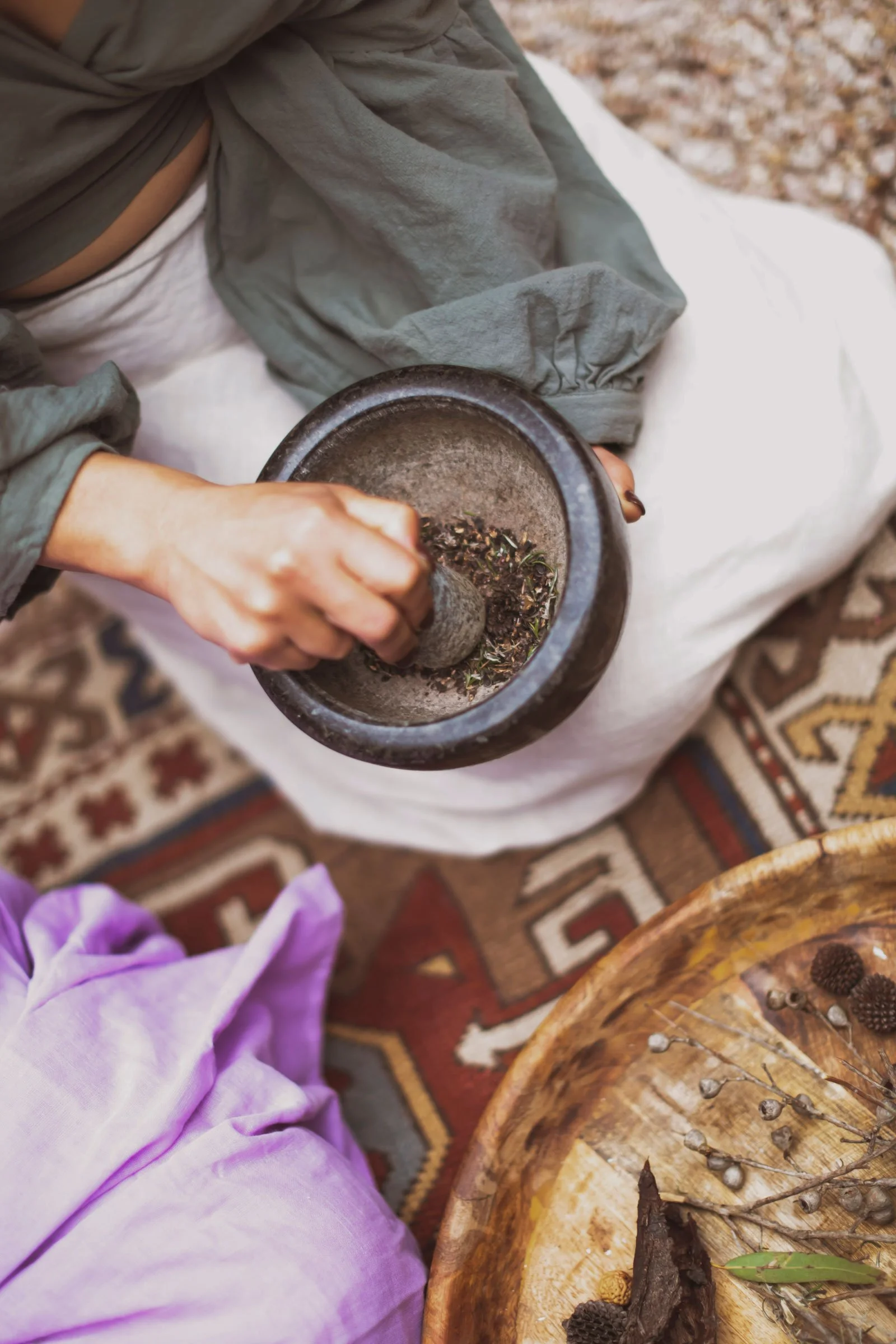 Person using a mortar and pestle to grind dried herbs or spices, seated on a patterned rug with a purple cloth and wooden tray with dried plants nearby.