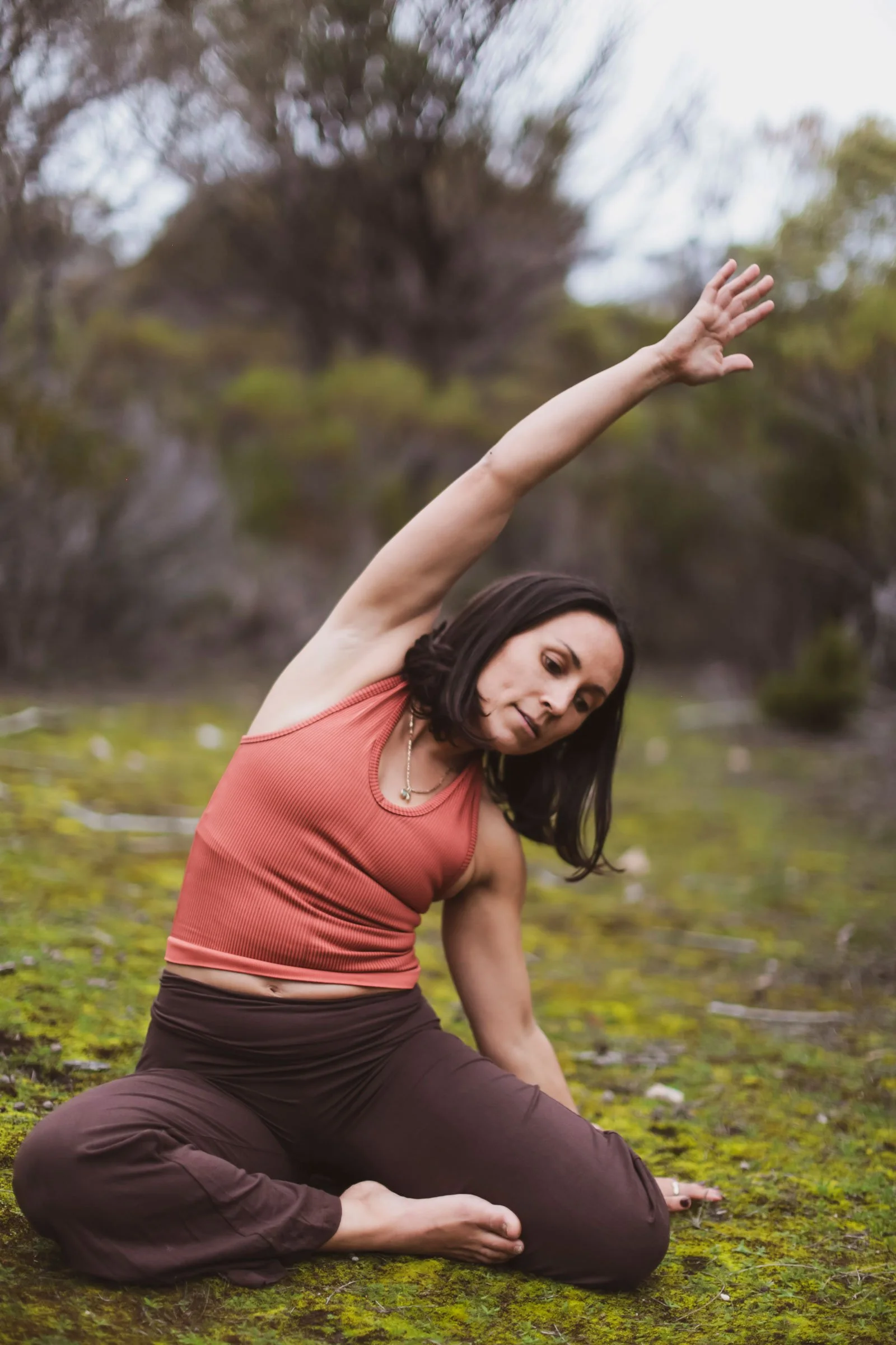 Woman practicing yoga outdoors on mossy ground, sitting with her legs crossed and stretching her right arm overhead, with trees in the background.