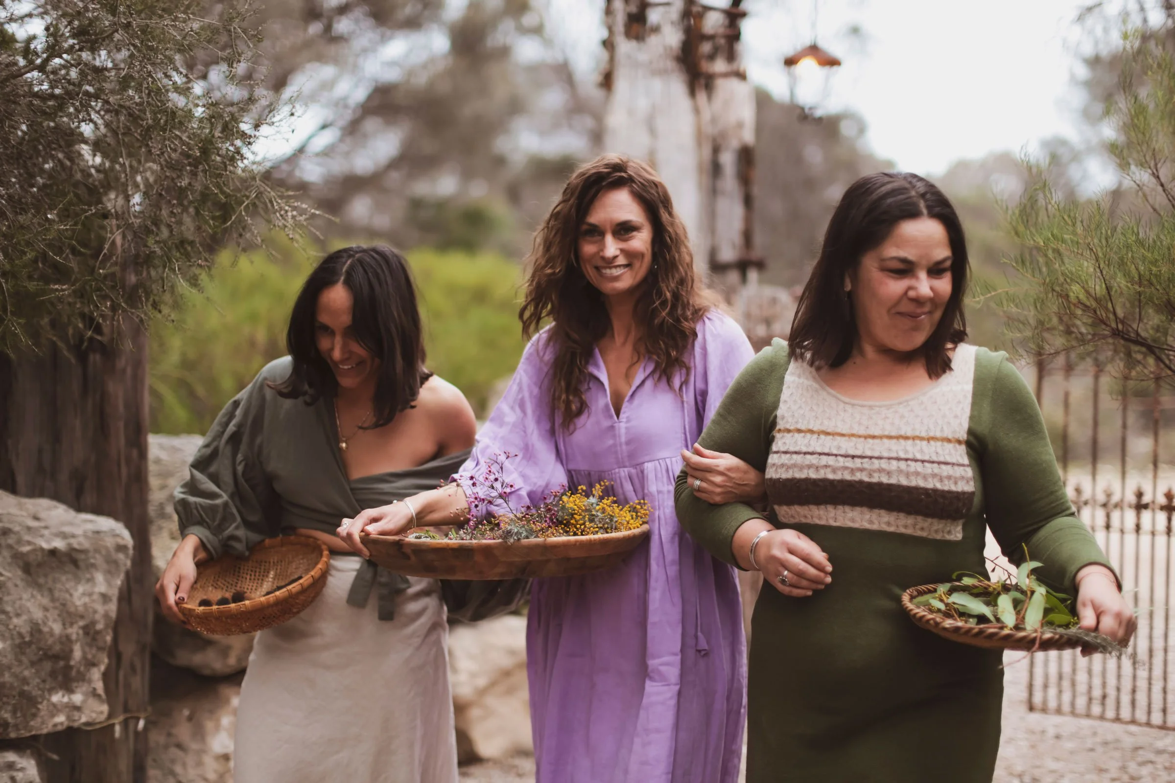 Three women walking outdoors, smiling, holding baskets with flowers and greenery, surrounded by trees and rocks.