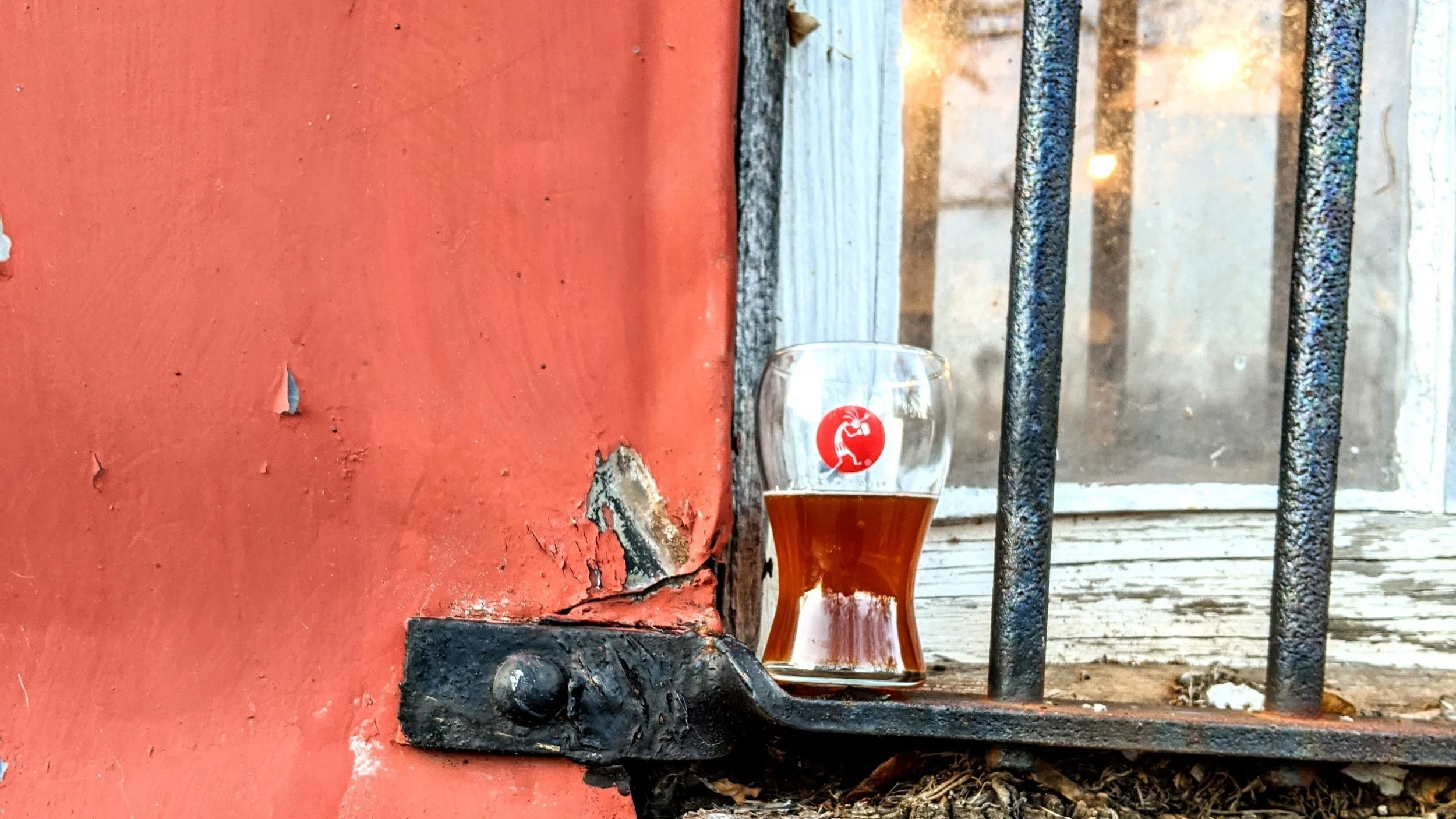 A glass of dark beer placed on a rusty metal rail outside a window with black metal bars and peeling red paint on a wooden wall. This brewery is located inside a  1900's feed and seed barn and often called the BrewBarn