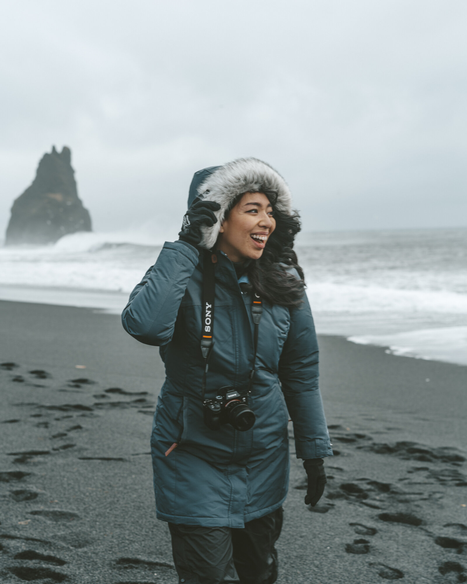 女孩微笑在黑色的沙滩上穿着冬季派克大衣，手套，和索尼a7相机