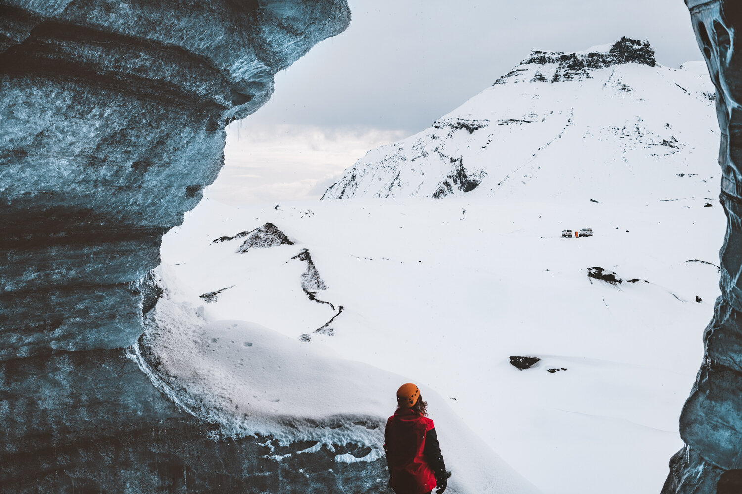 一个穿着红色夹克、戴着橙色头盔的小人从一个冰冷的冰川洞穴里凝视着山上的雪景