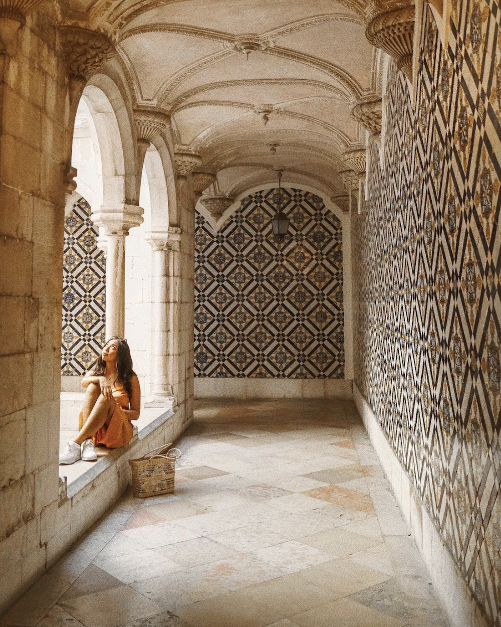 brunette woman in gold-orange dress sits in a tiled courtyard of white arches and blue detail