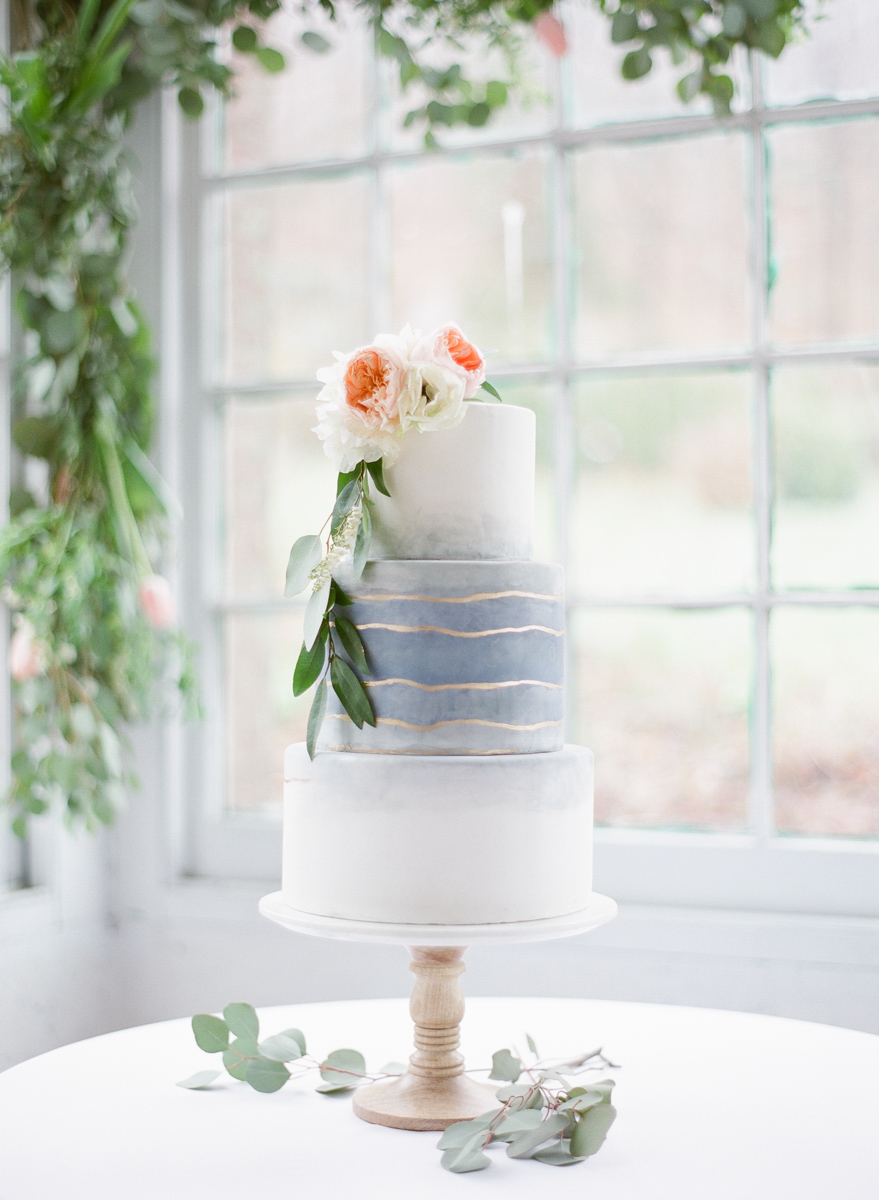 As Spring inspires fresh bright blooms and warm light, our wedding shoot took on these lovely spring accents and soft painterly details. Shot at the stunning Lace Factory in Deep River, Connecticut, the interior of this historic industrial building provided a captivating juxtaposition to the ethereal look of our bride and organic styling of the table arrangement. In addition to the atmosphere of the factory, I hand painted a backdrop with shades of blue to compliment our bride’s beautiful red hair. All the details were styled to feel effortless and timeless with touches of pale pink accented against the blue. Loose flowing ribbons adorned the romantic unstructured bridal bouquets handcrafted by Blush Floral Design . Our farm table lush with blooming tulips and white candles was topped by a unique hanging floral center piece. Flowing watercolor elements are found in the invitation design by Tanya Costigan Events and in the heavenly painted detail of the cake by Erica O’Brien Cake Design .