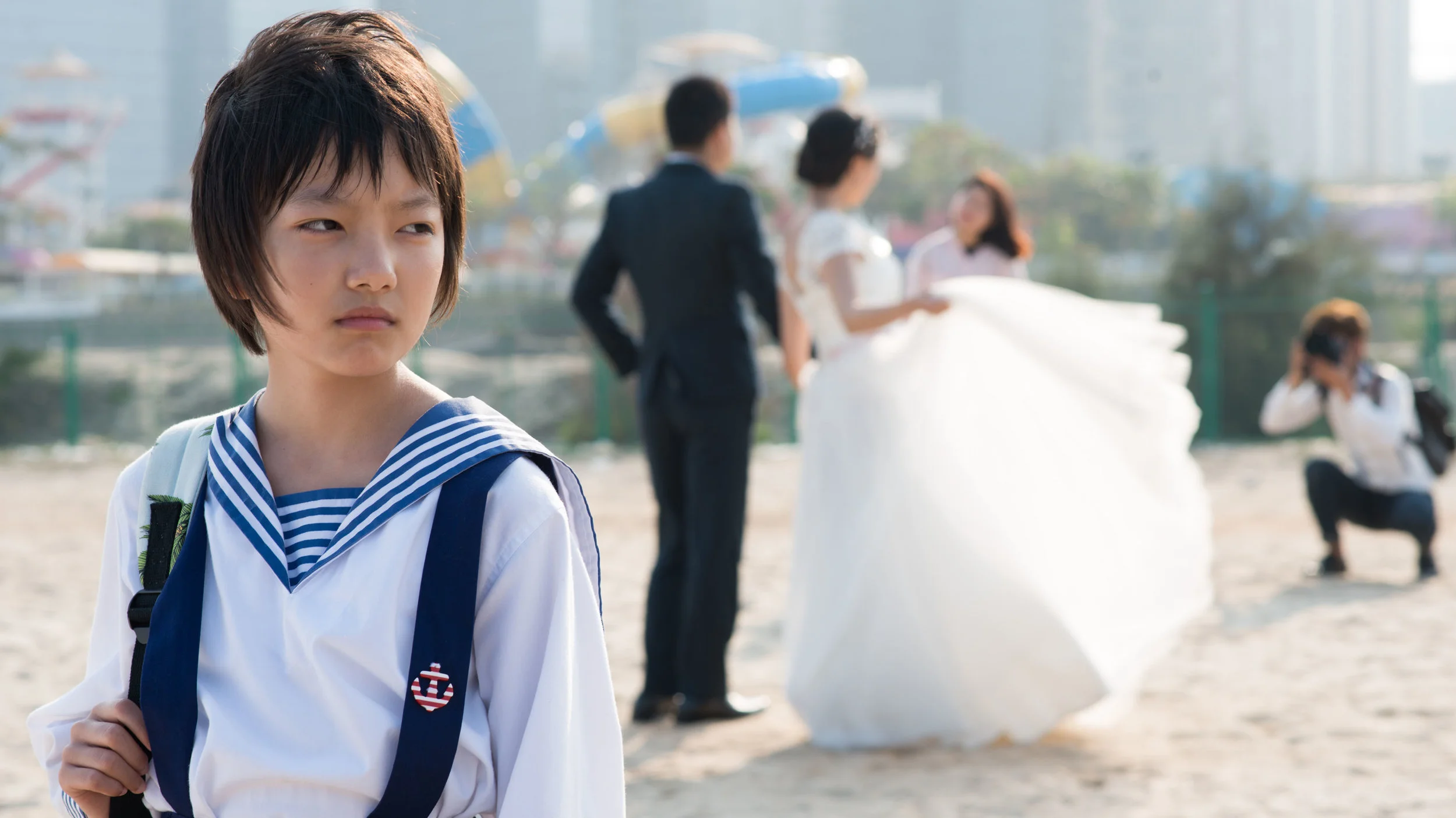 Traumatized Wen (Meijun Zhou) surrounded by happy tourists taking romantic wedding pictures on the beach.