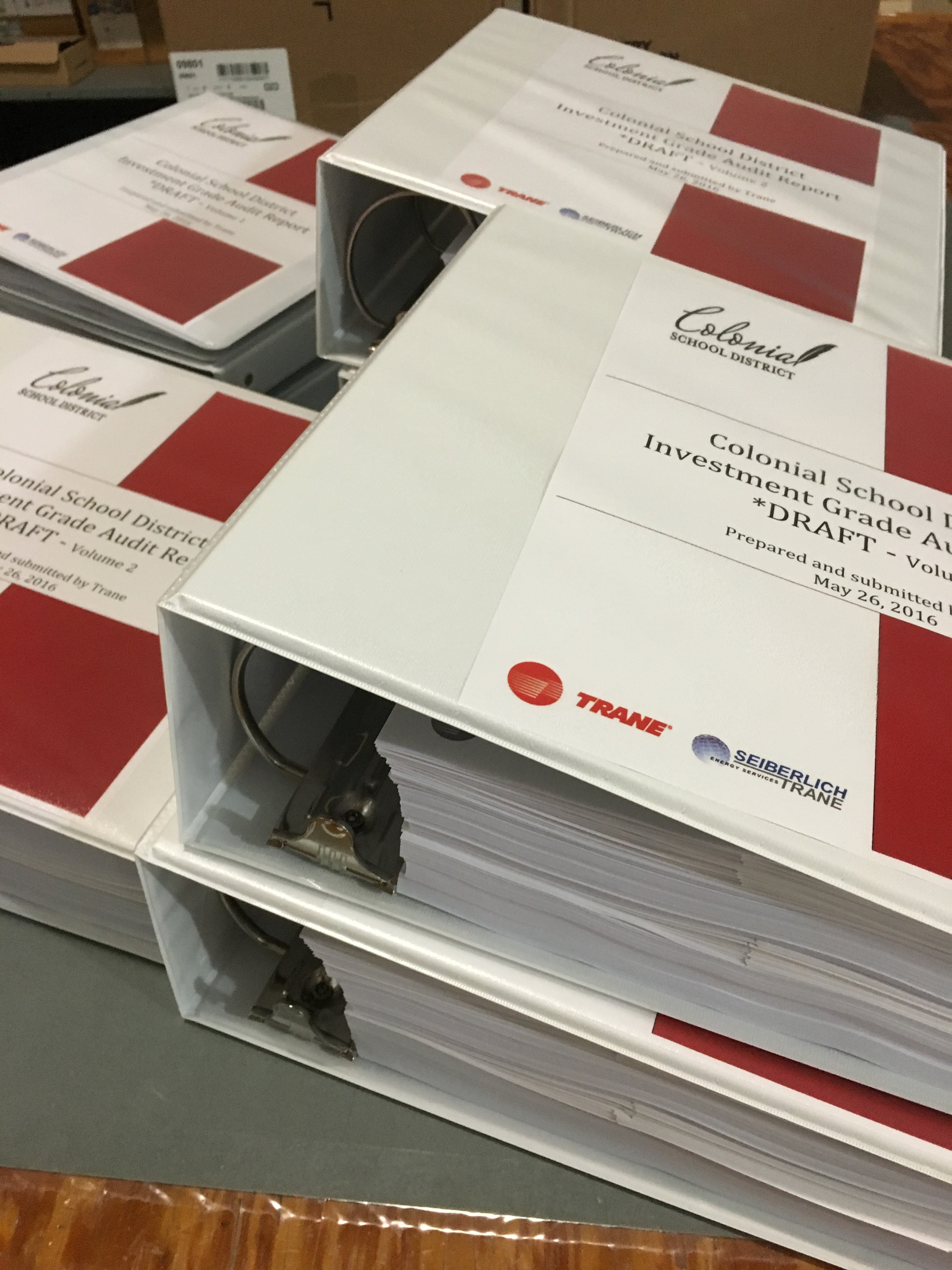 Two white binders with papers inside, labeled Colonial School District Investment Grade Audit Draft, Volume 2, with the Trane and Seiberlich Energy Services logos, on a gray surface.
