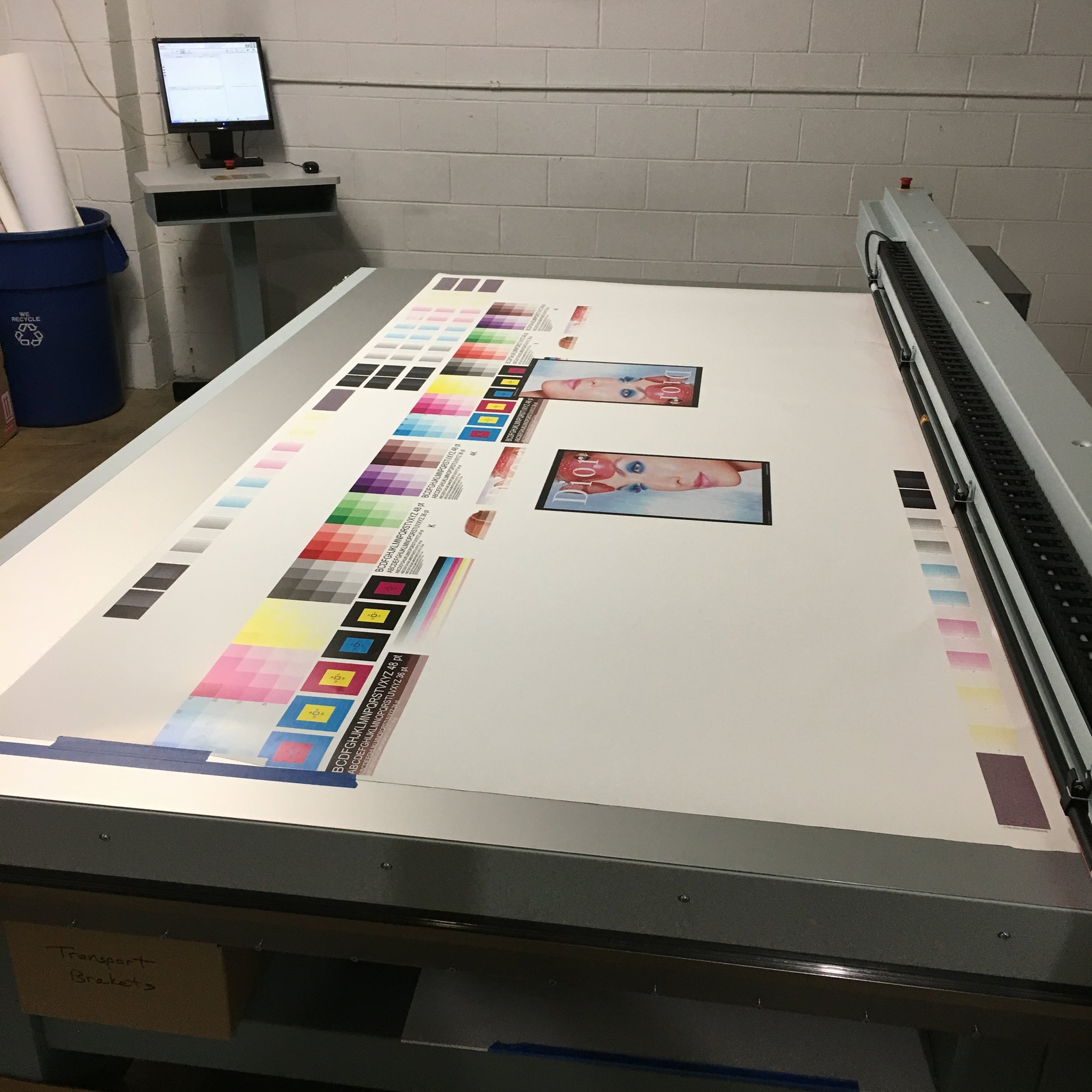 Large-format printer printing a magazine cover featuring a woman's face with blue makeup and red hair. Color calibration strips and test images are on the print bed. A computer monitor is visible in the background.