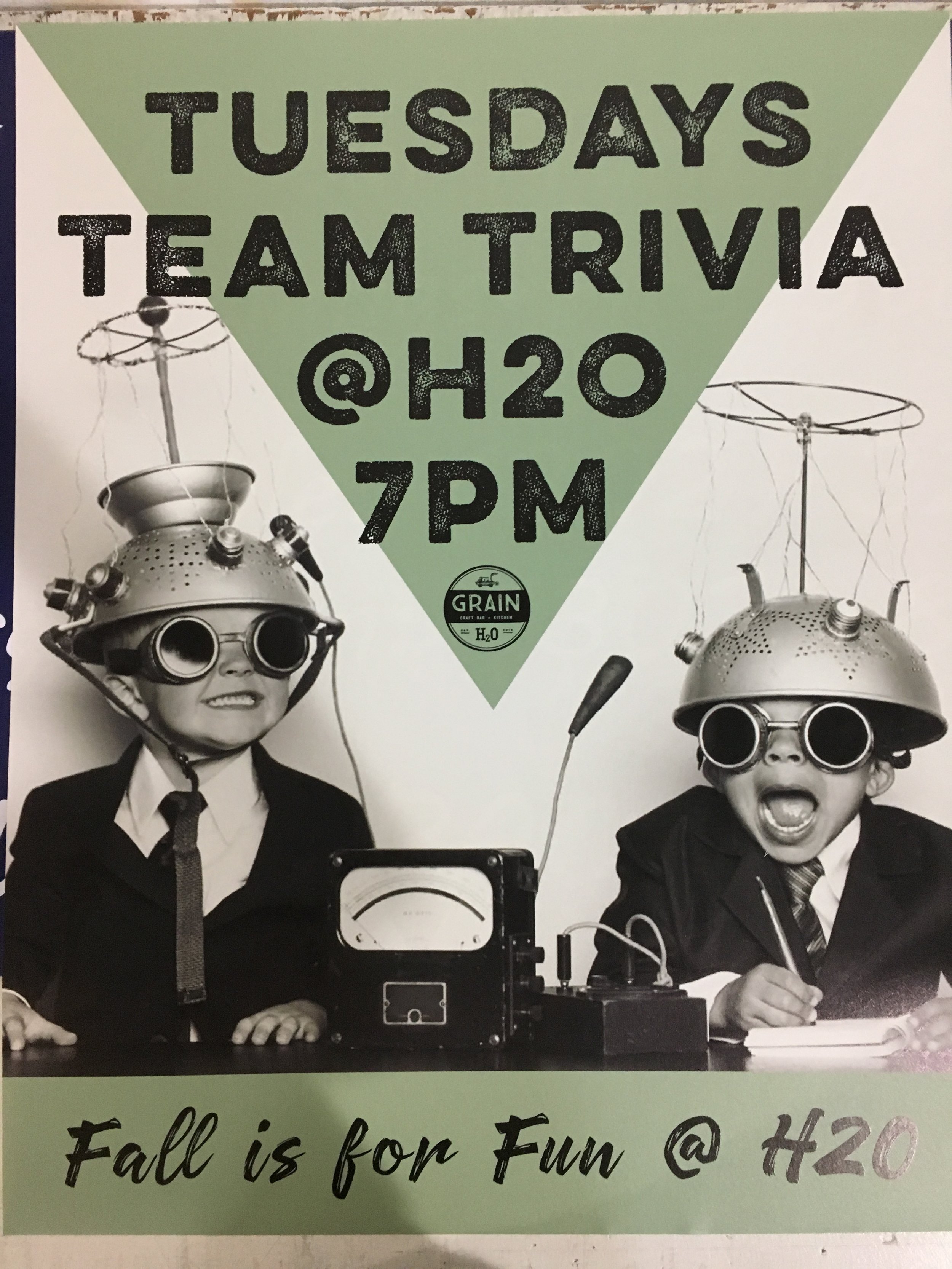 Black and white photo of two children dressed as scientists with vintage headgear and goggles, sitting at a desk with a microphone and a gauge, promoting Tuesday night team trivia at H2O at 7 PM.