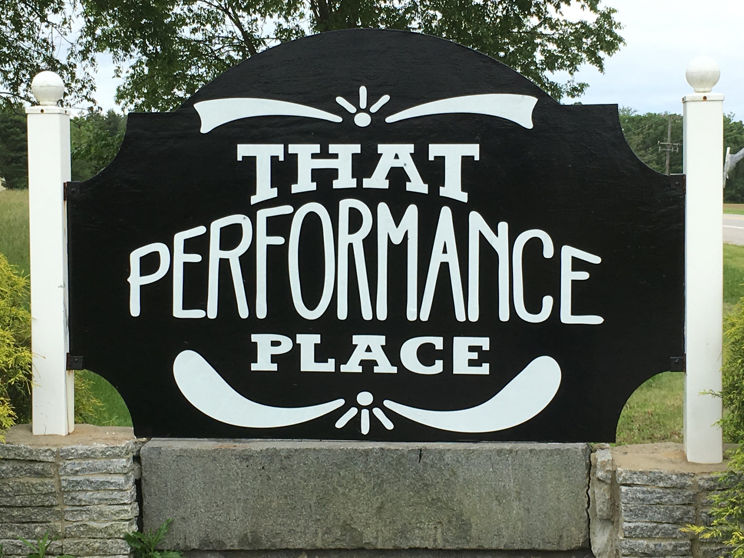 Black outdoor sign with white lettering that reads 'That Performance Place' and decorative white accents, mounted on stone and wood posts, in a natural outdoor setting.