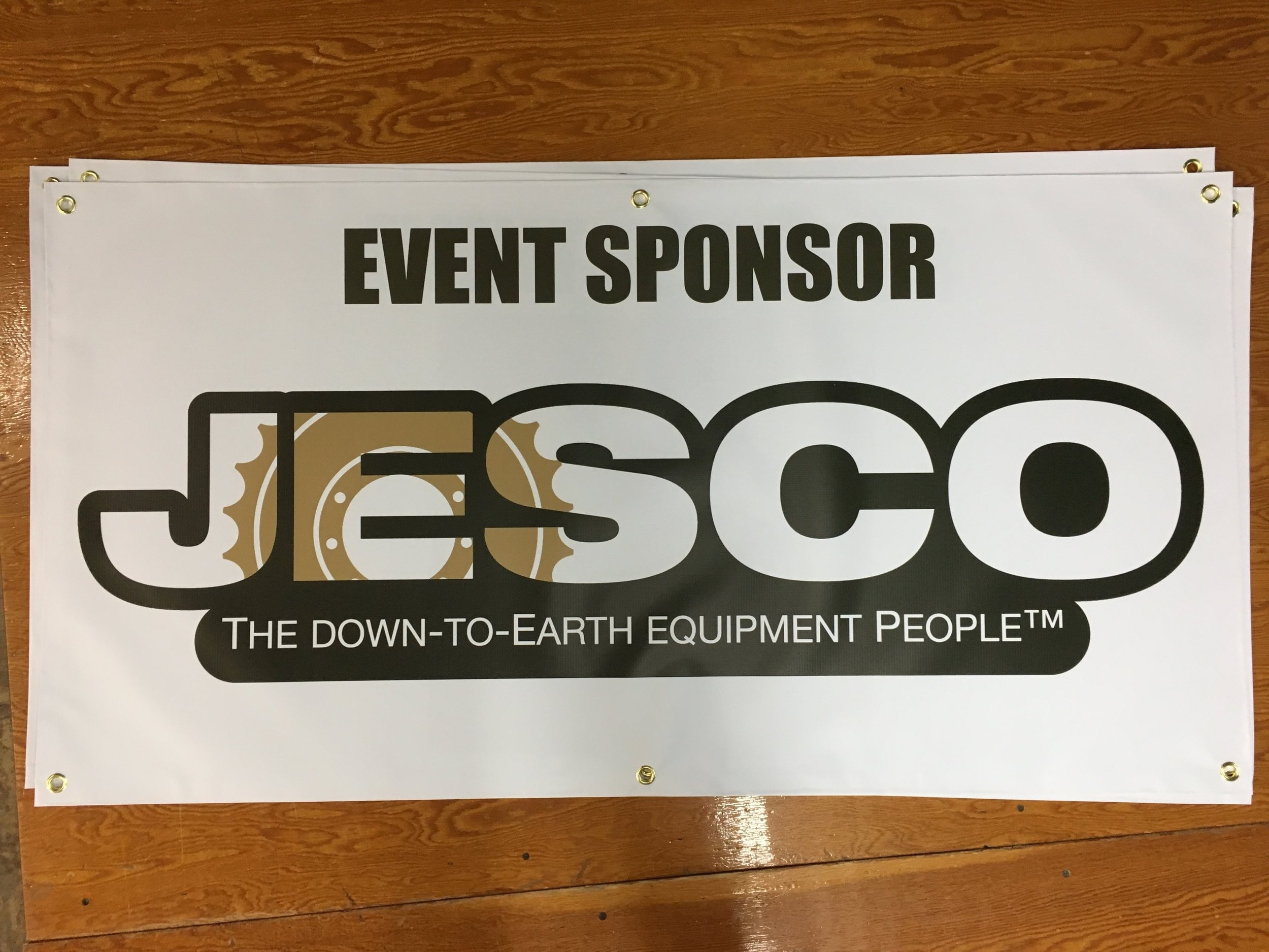 Sign with event sponsor information featuring the logo of JESCO and the slogan 'The Down-to-Earth Equipment People' on a wooden surface.