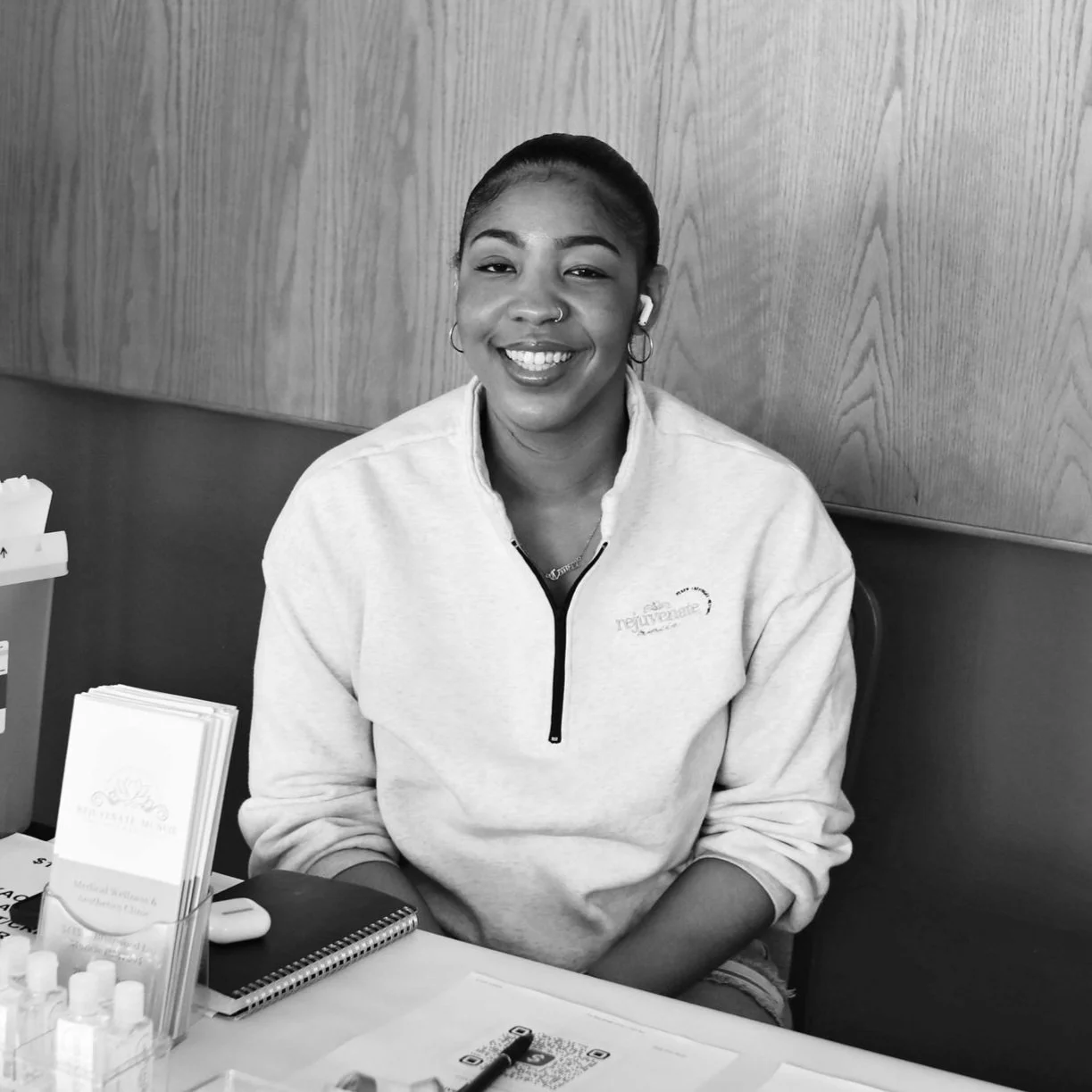 A smiling woman with dark hair in a bun, wearing a light-colored zip-up sweatshirt, sitting at a table with promotional materials and small bottles.
