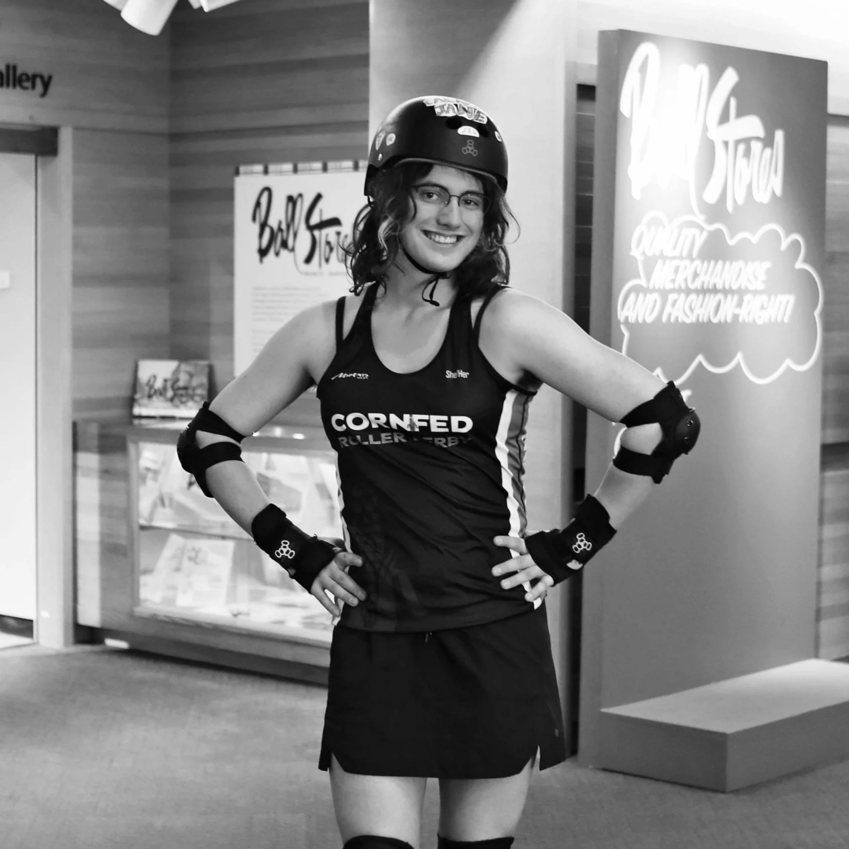 Young woman dressed in roller derby gear, smiling, standing indoors in front of a display with the text "Ball Stop" and a sign that reads "Quality merchandise and fashion-right."