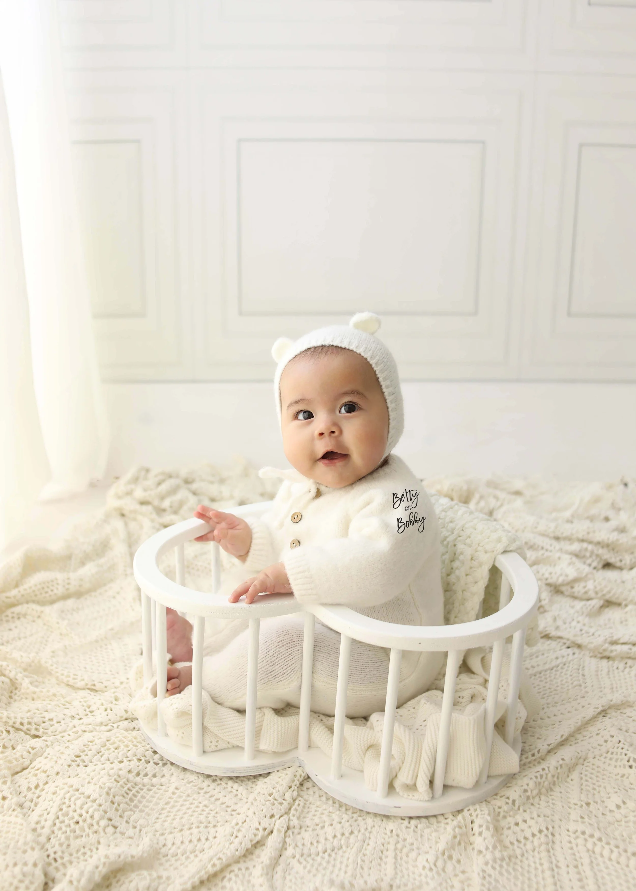 A baby sitting inside a white wooden cradle on a cozy knitted blanket, wearing a cream onesie and matching knit hat with bear ears, in a bright room with white walls.