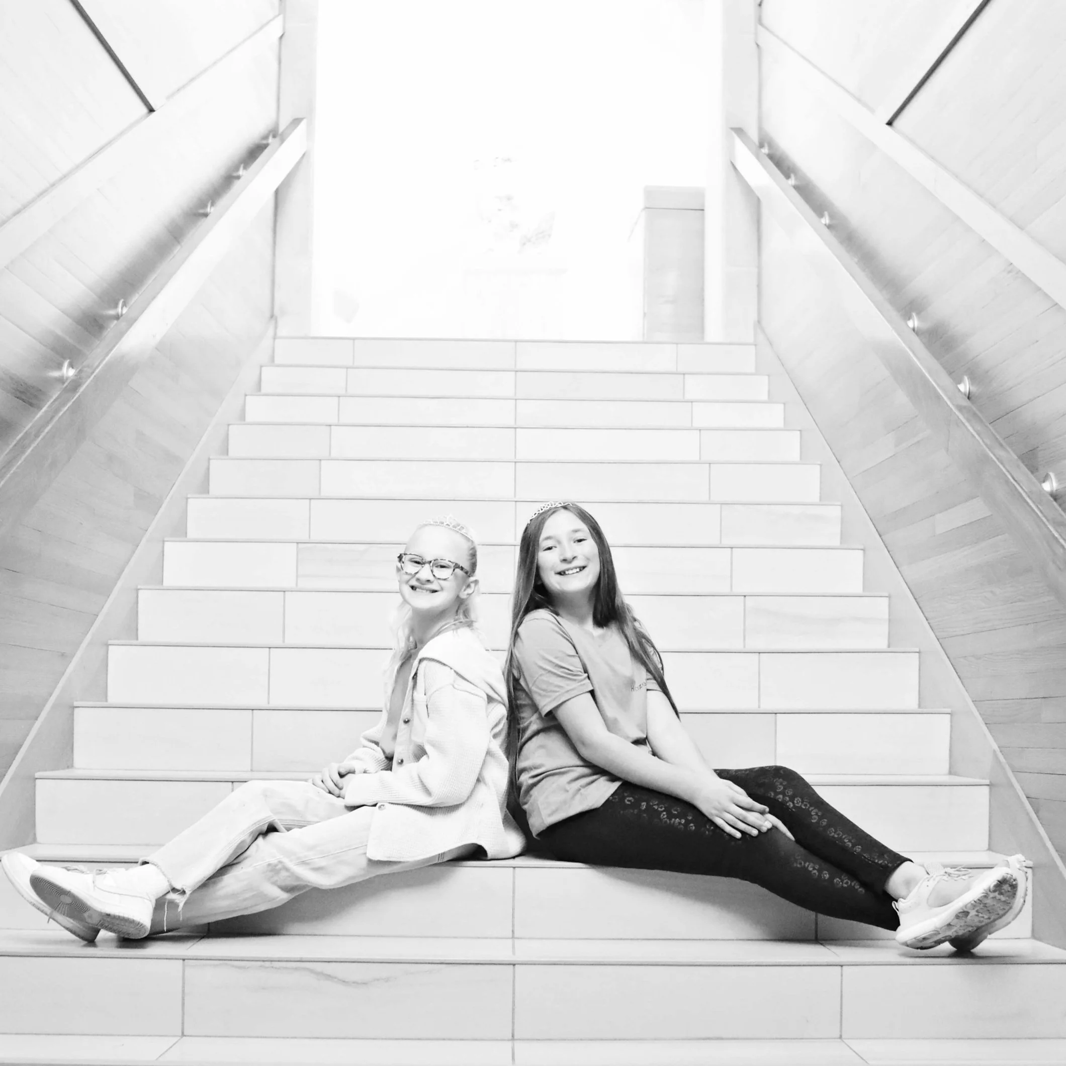 Two young girls sitting on the stairs, back to back, smiling, in an indoor setting.
