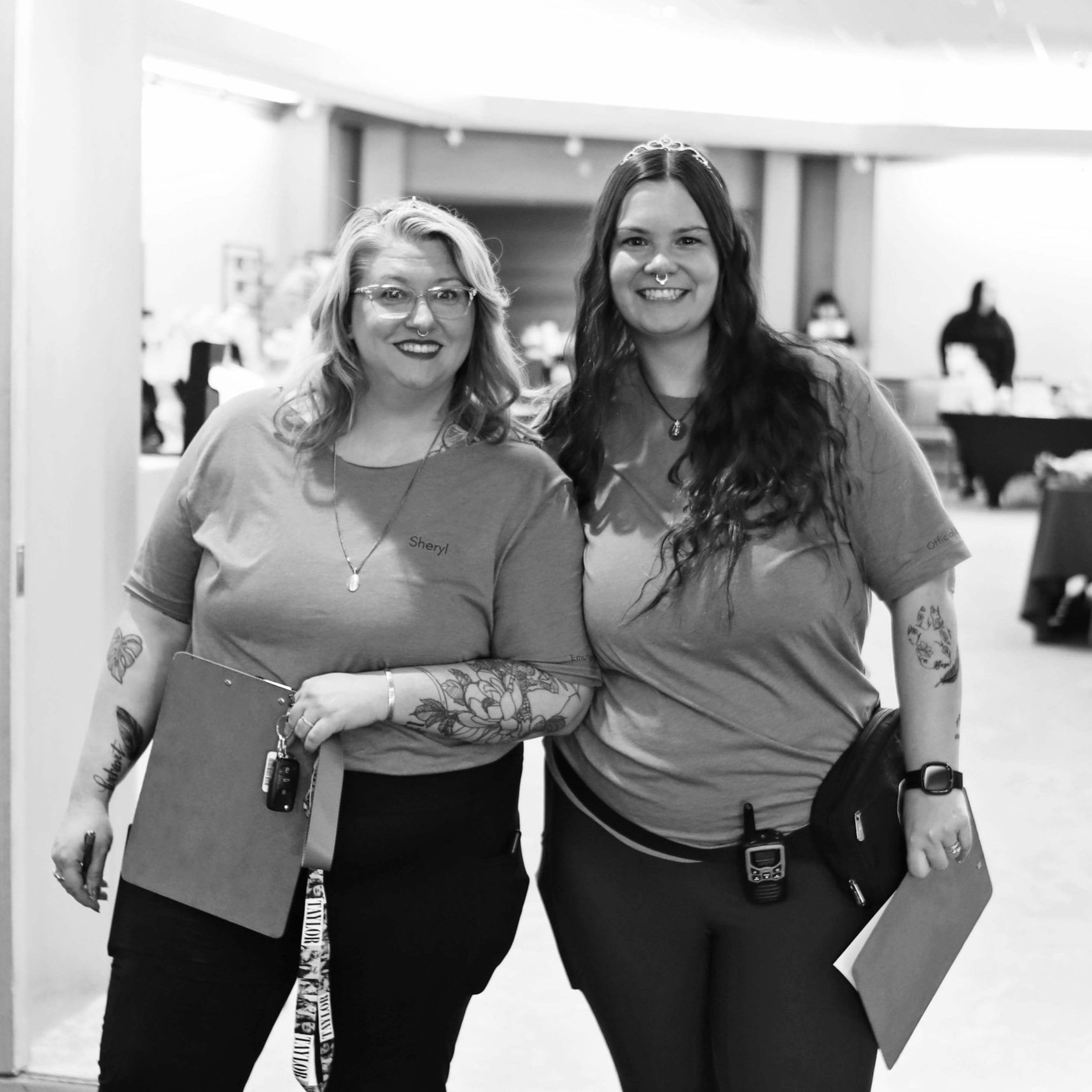 Two women standing side by side indoors, smiling at the camera, wearing casual shirts with name tags, holding clipboards, and carrying walkie-talkies, in a setting with tables and people in the background.