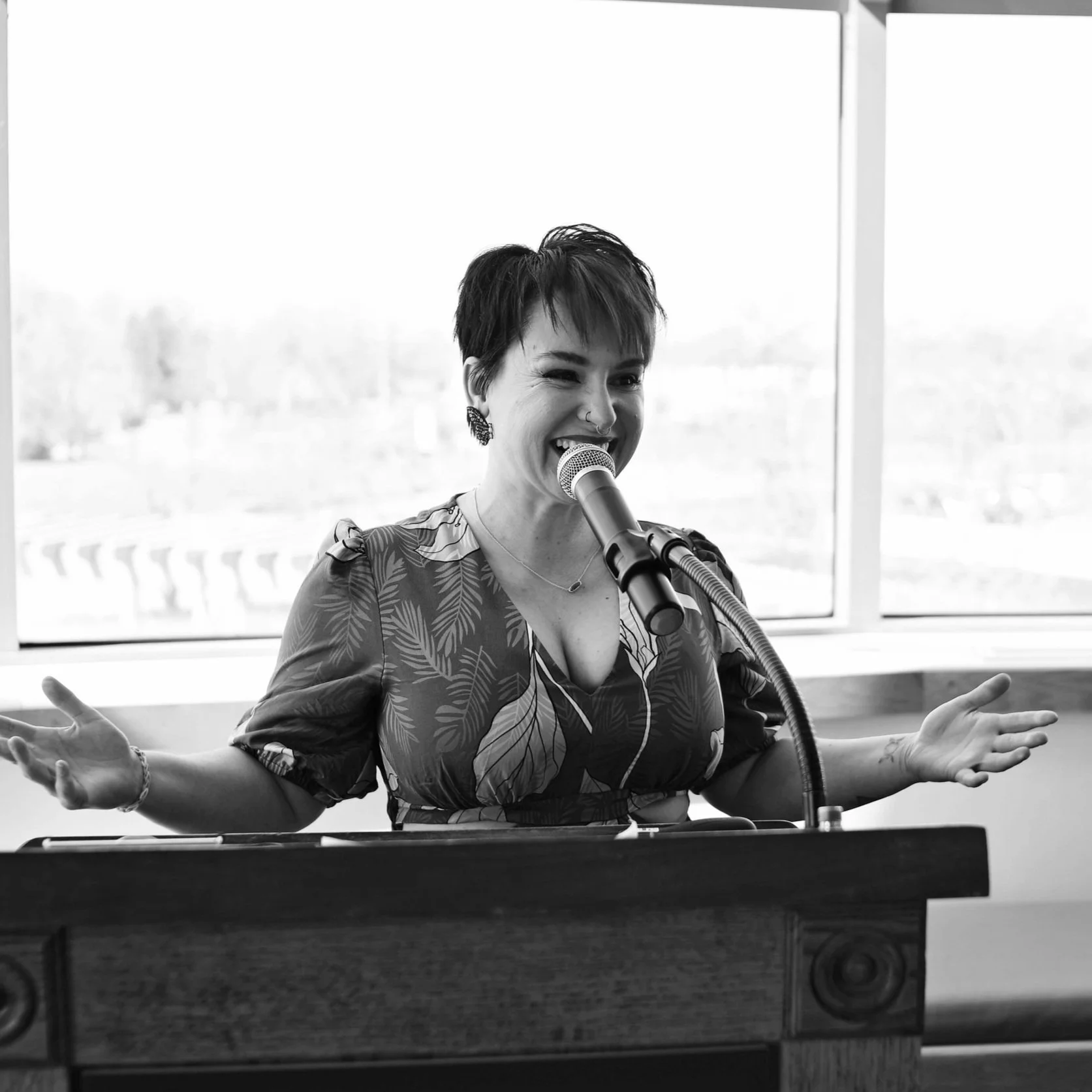 Black and white photo of a woman with short hair and earrings speaking into a microphone, standing behind a podium in front of a window.