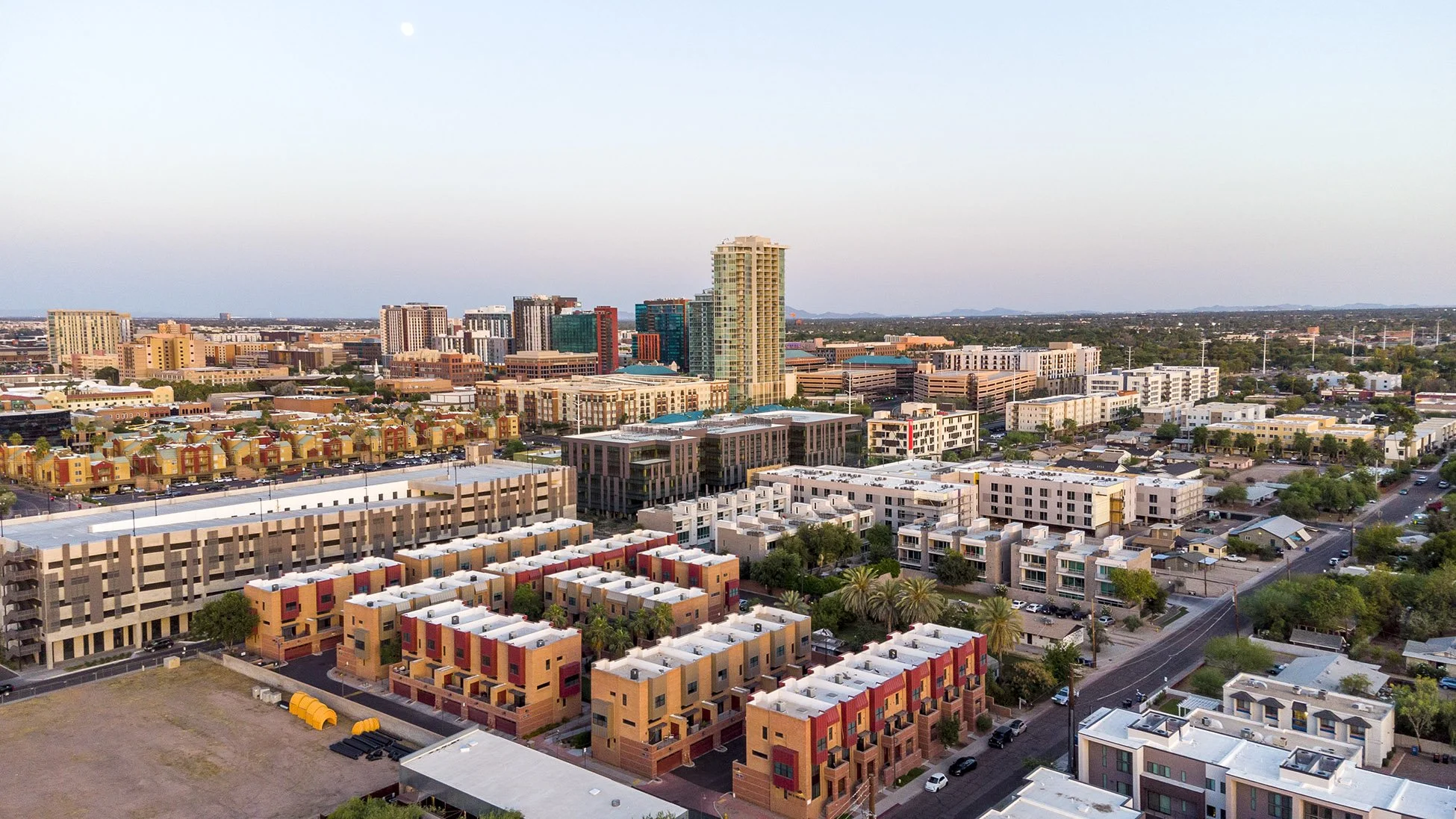 Aerial view of Tempe, Arizona with a modern city skyline with tall buildings, mid-rise apartments, and colorful residential houses under a clear sky with the moon visible.