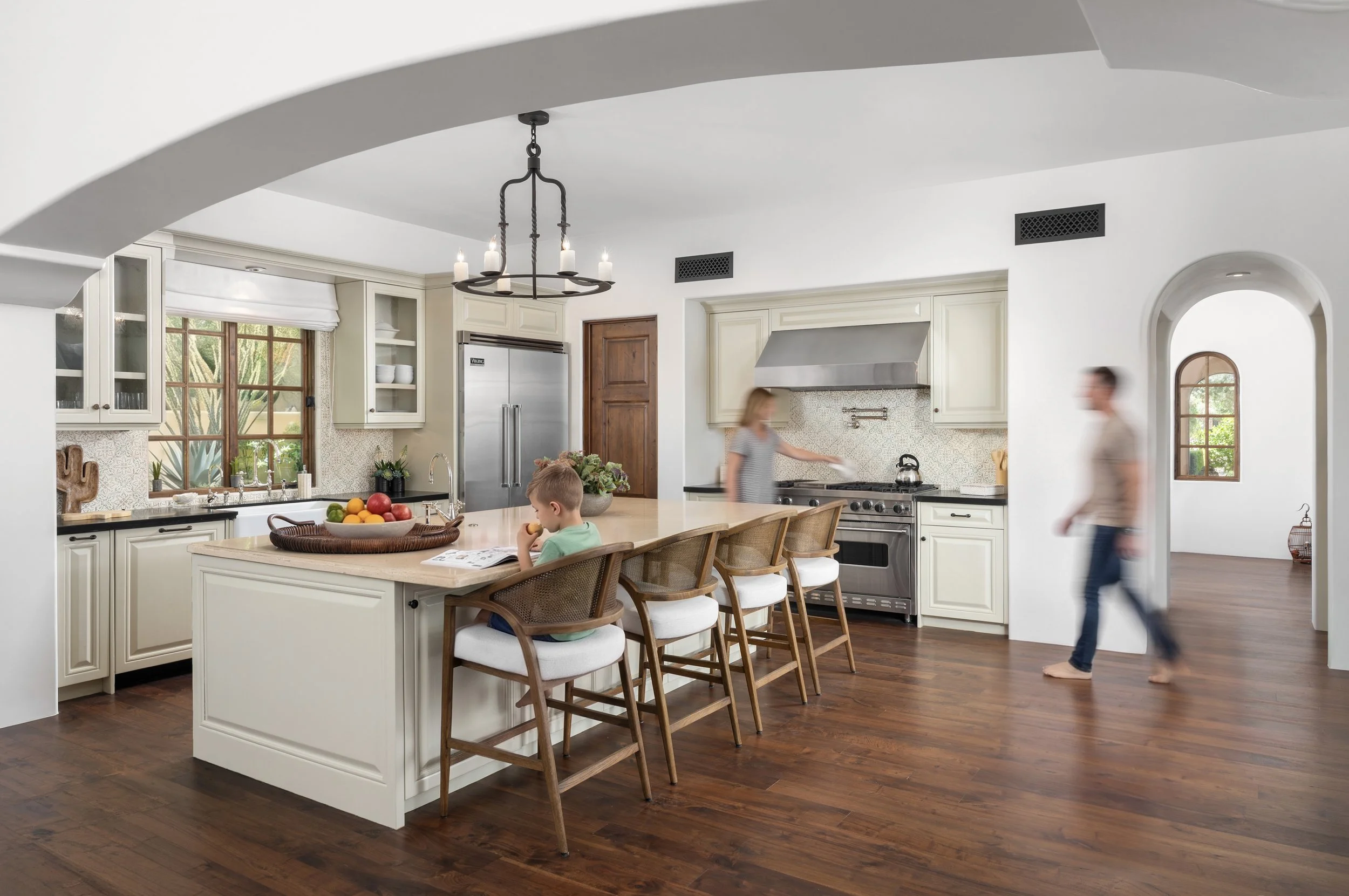 A spacious kitchen with a large island, cream-colored cabinets, stainless steel appliances, a chandelier hanging from the ceiling, hardwood floors, and two children sitting at the island while an adult walks by in the background.