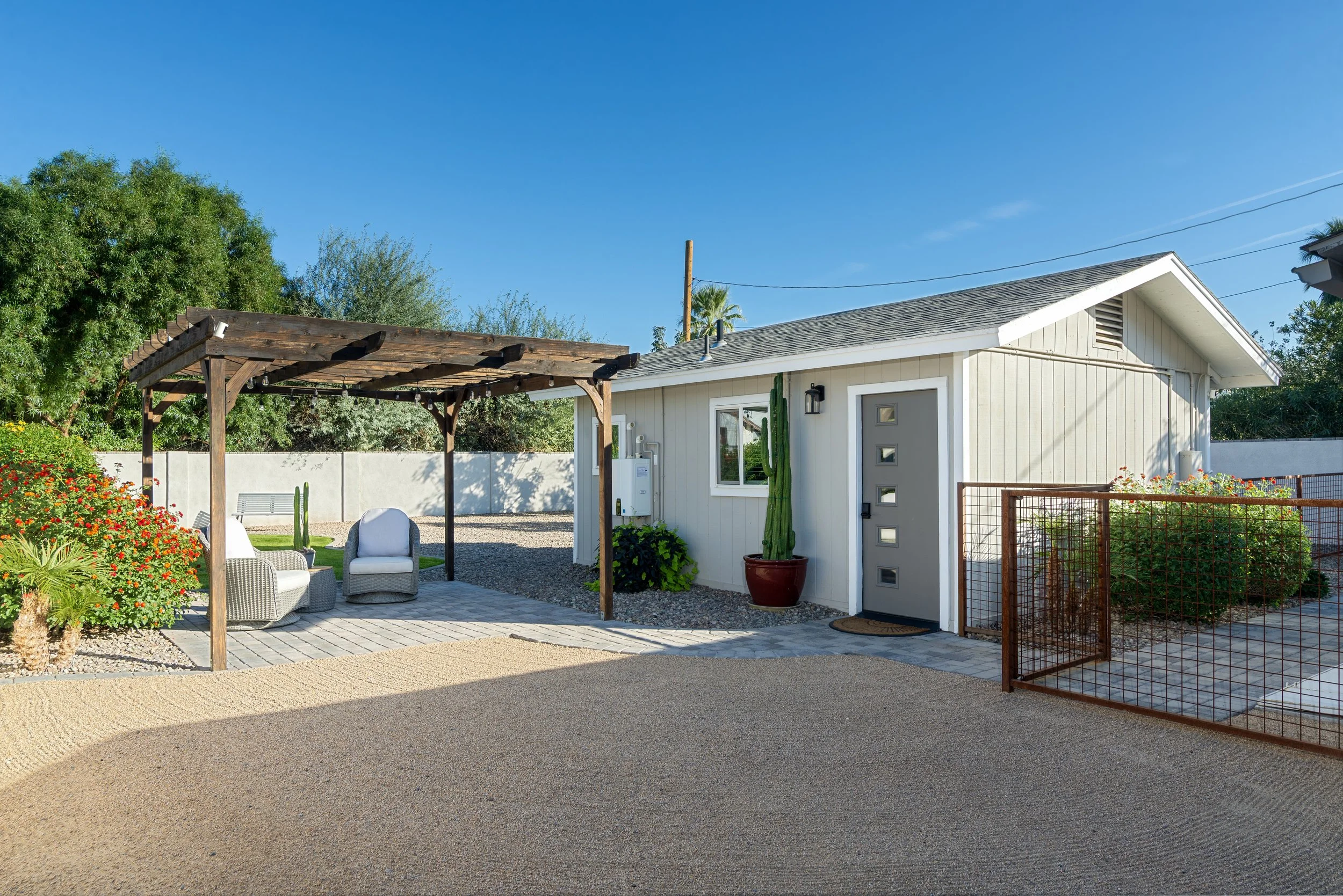A backyard patio with a wooden pergola, outdoor chairs, a small table, lush greenery, a white house, and a gravel pathway under a clear blue sky.
