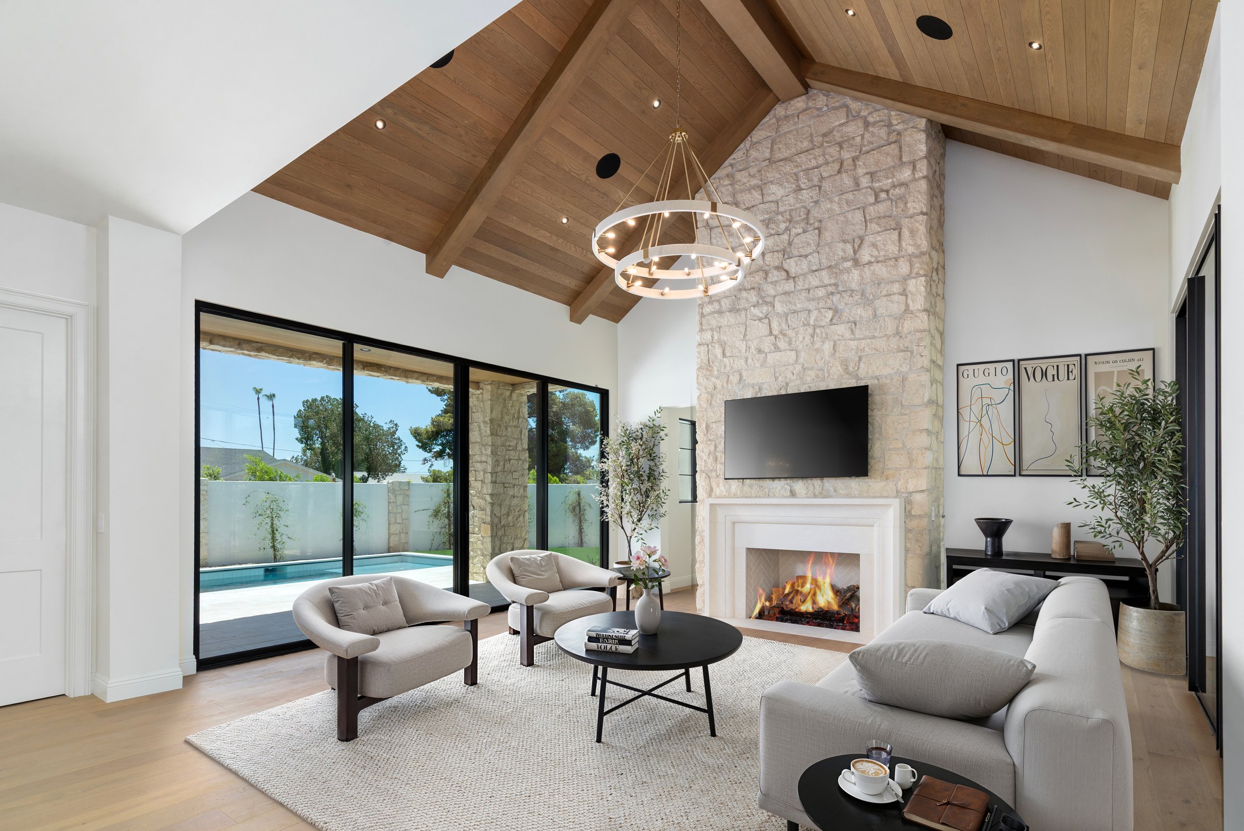 Modern living room with a vaulted wooden ceiling, stone fireplace, large window with a view of a backyard pool, and stylish furniture including white sofas, chairs, and a black coffee table.