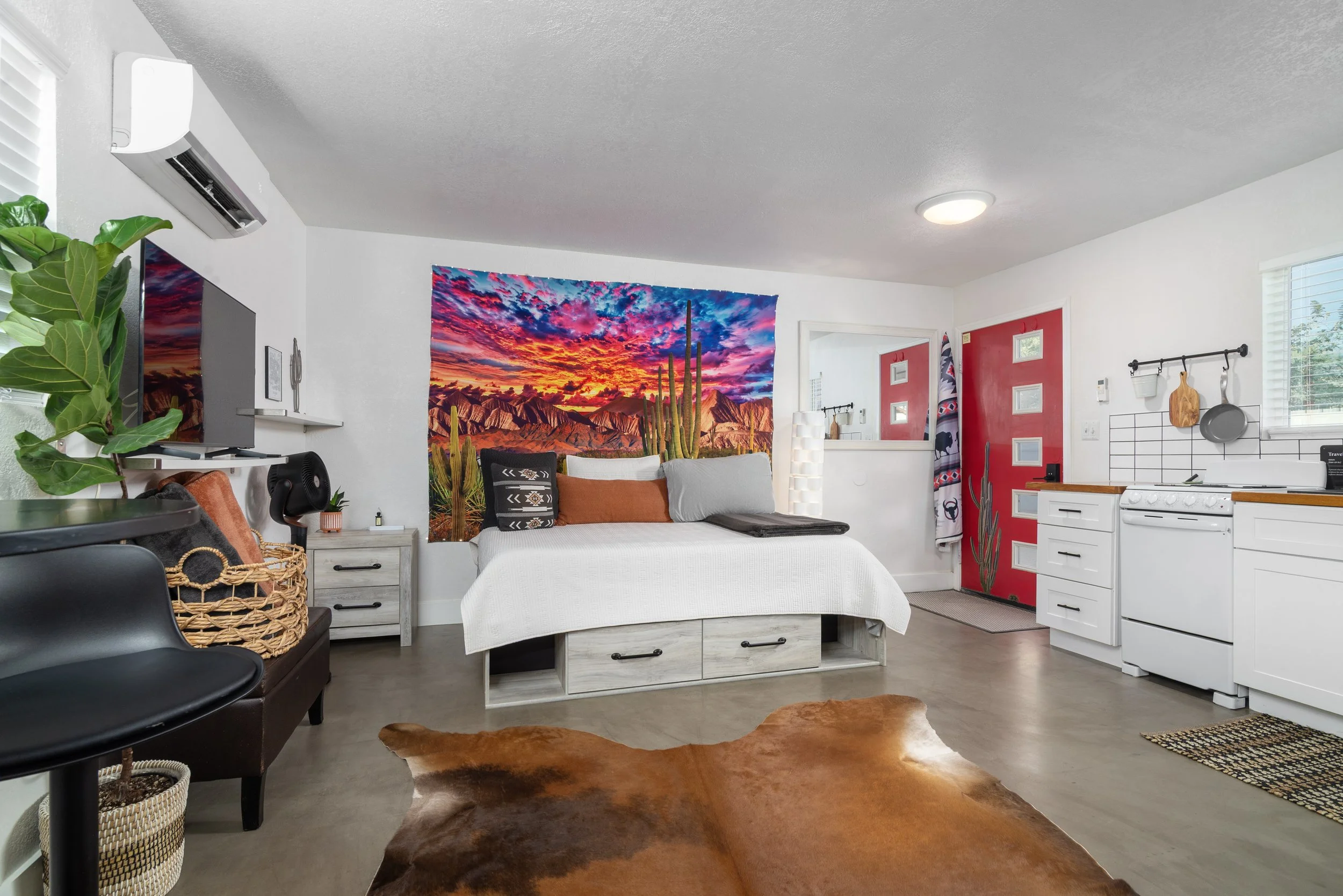 A cozy living room with a white couch, pillows, and a colorful desert sunset artwork on the wall. The room has plants, a cowhide rug, and kitchen cabinets with red doors.