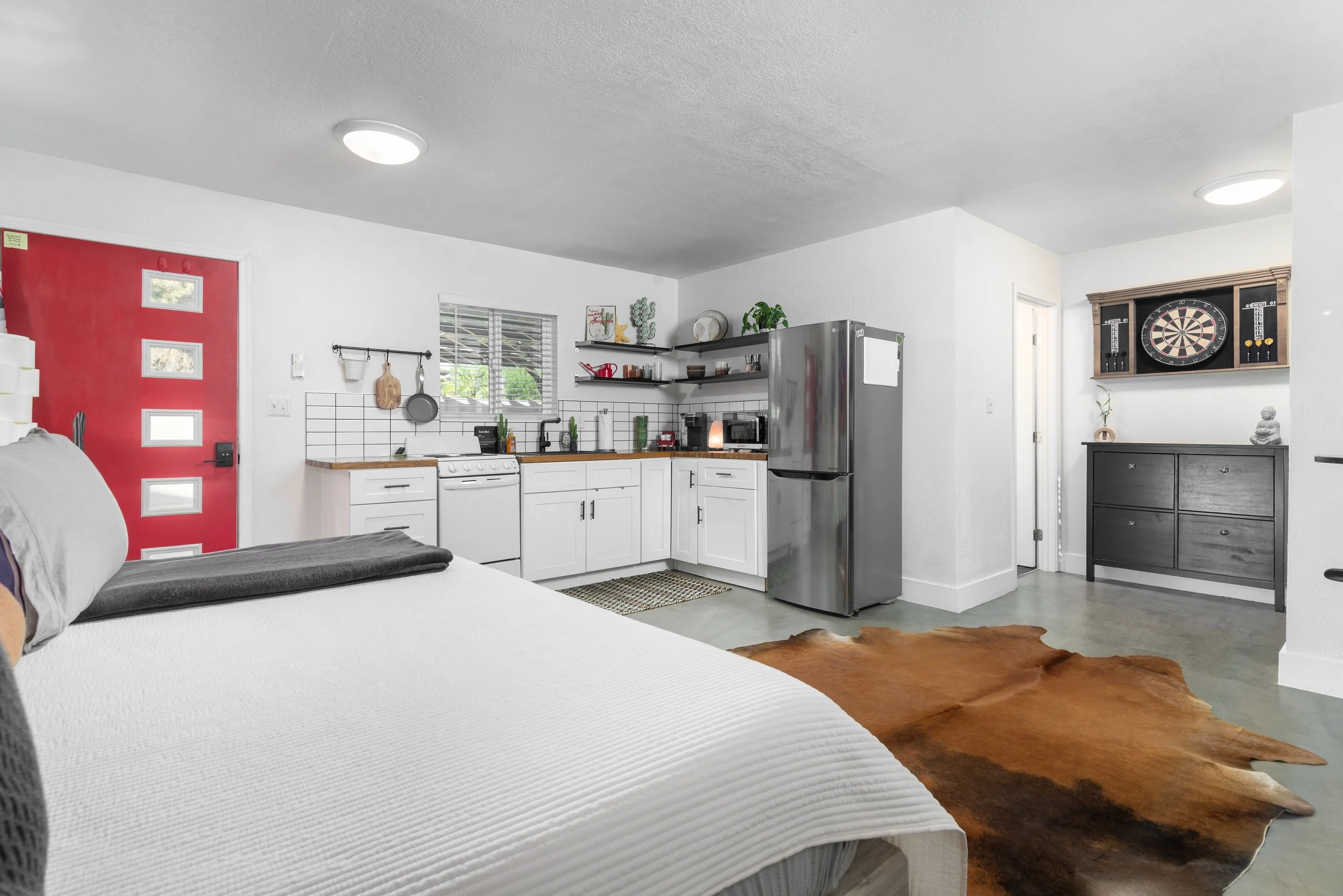 Interior of a small studio apartment with a partial view of a bed, kitchen area, and a dartboard on the wall.