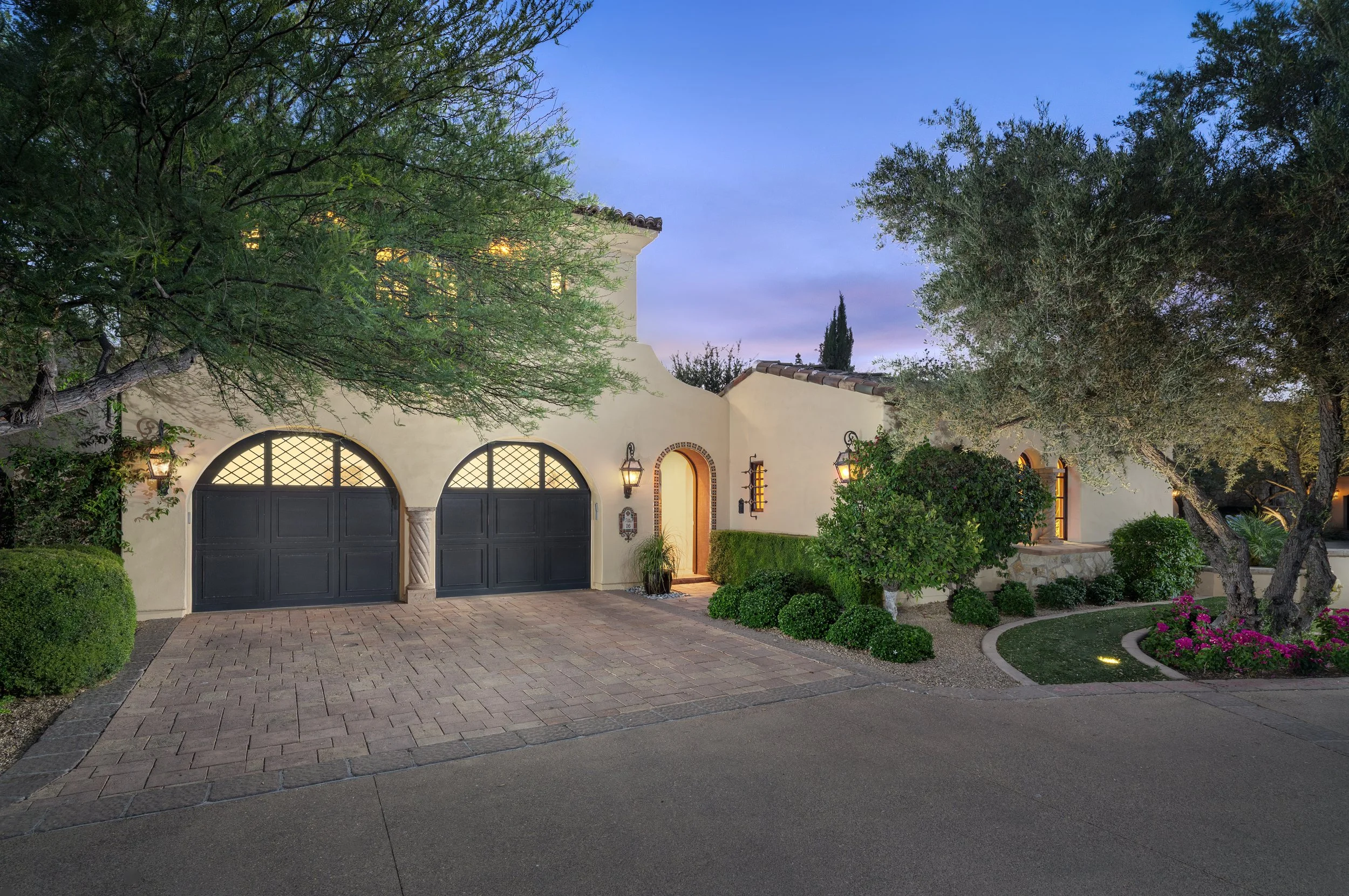 A beautiful two-story house with a stucco exterior and a tiled roof, surrounded by lush greenery and trees. The house features arched windows and doors, with warm outdoor lighting illuminating the entrance. The driveway is paved with bricks and bordered by neatly trimmed bushes and flowering plants, creating a welcoming and elegant ambiance during dusk.