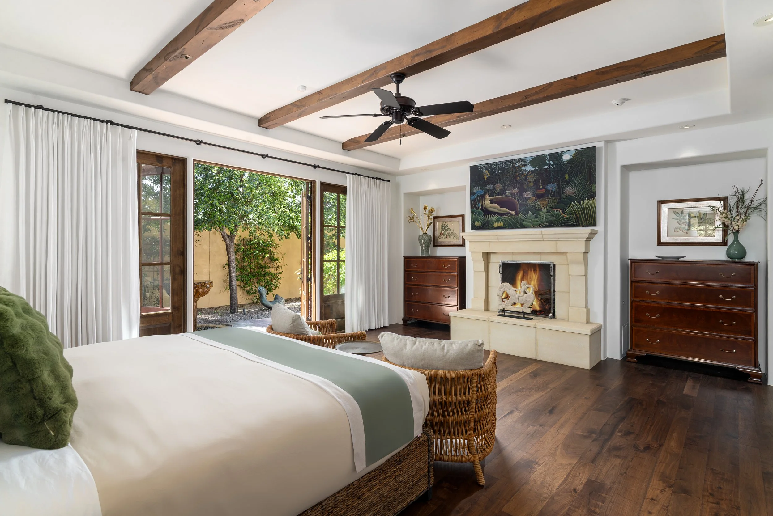 Living room with open glass doors leading outside to a garden, white curtains, wooden ceiling beams, a fireplace with a fire, and dark wooden furniture, including dressers and decorative items.
