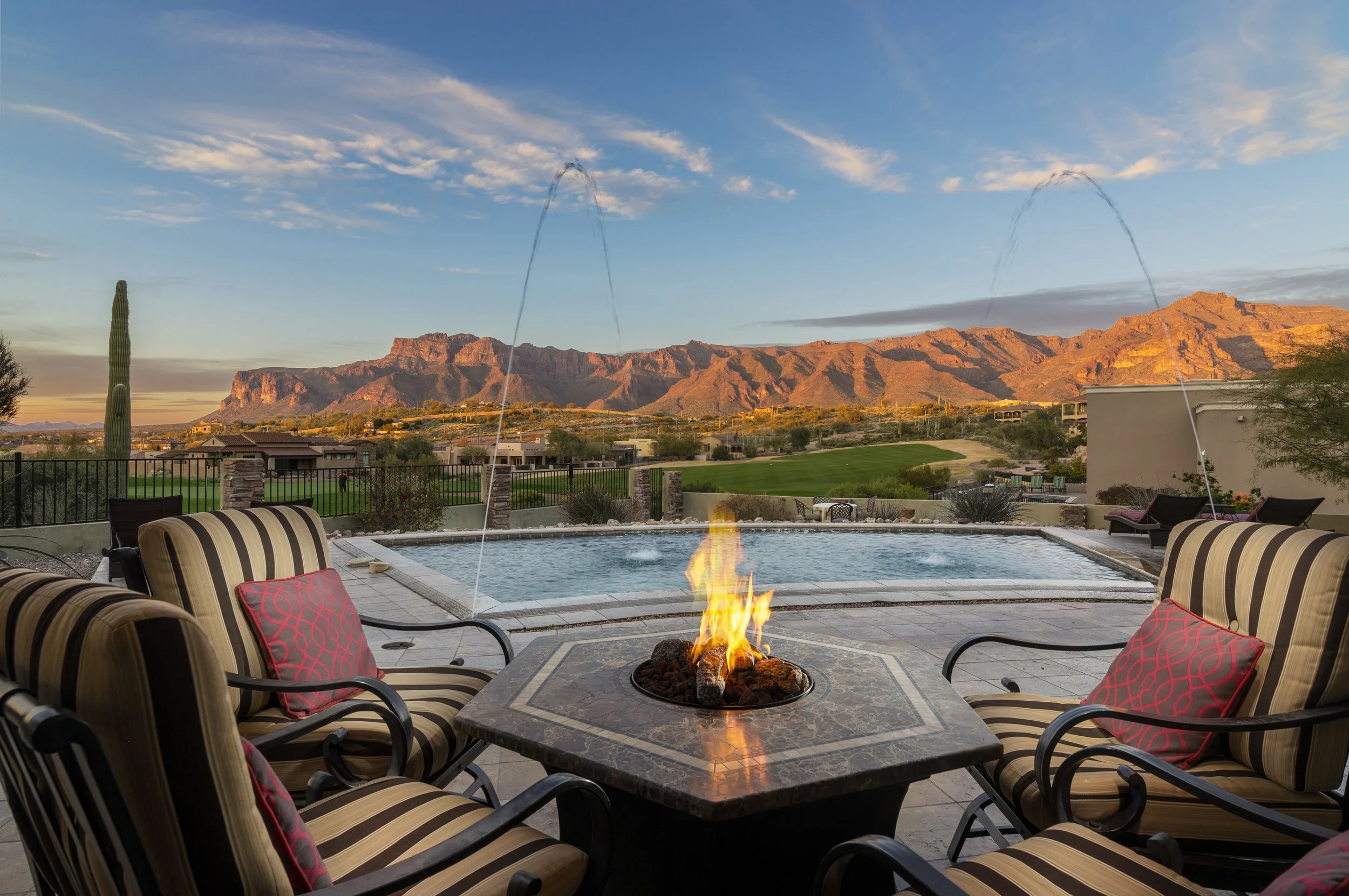 Outdoor patio with a fire pit, striped chairs with red pillows, swimming pool, and a mountain range in the background at sunset.